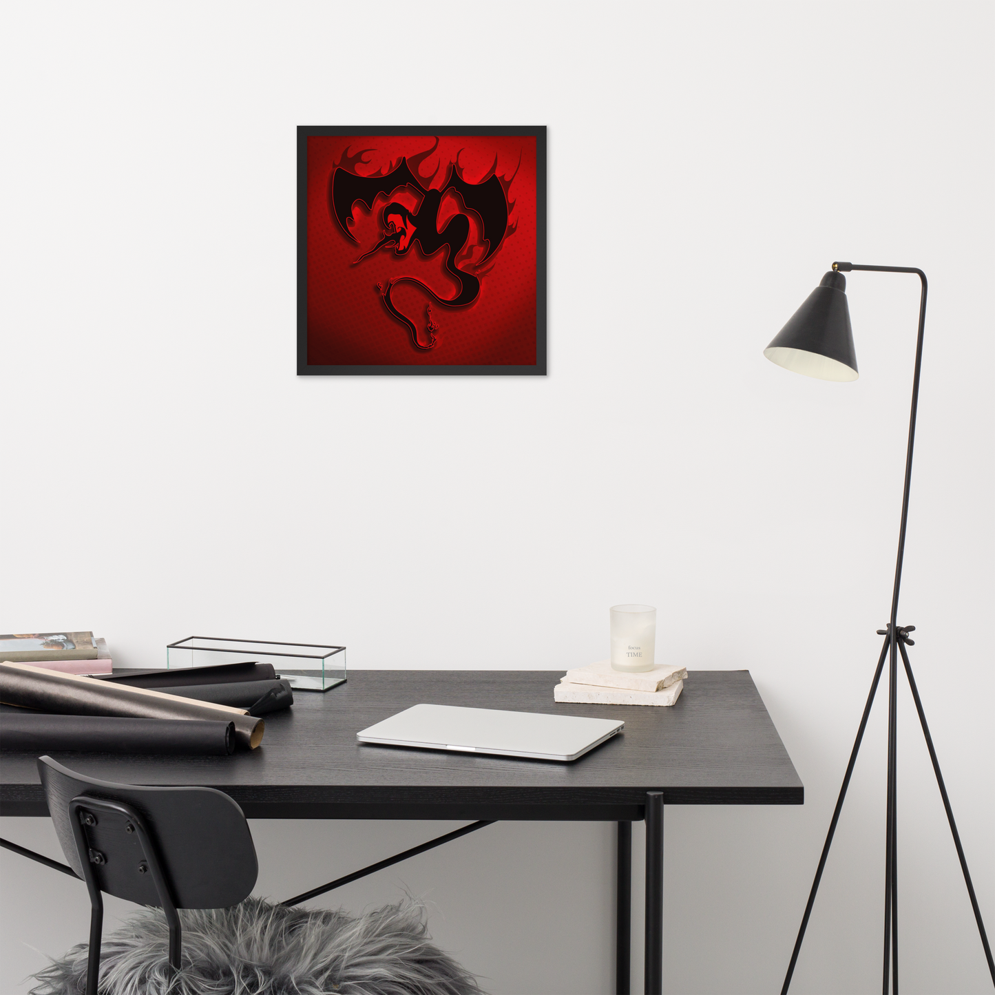 Framed red firebreathing winged snake artwork on a wall above a desk with a lamp and books.