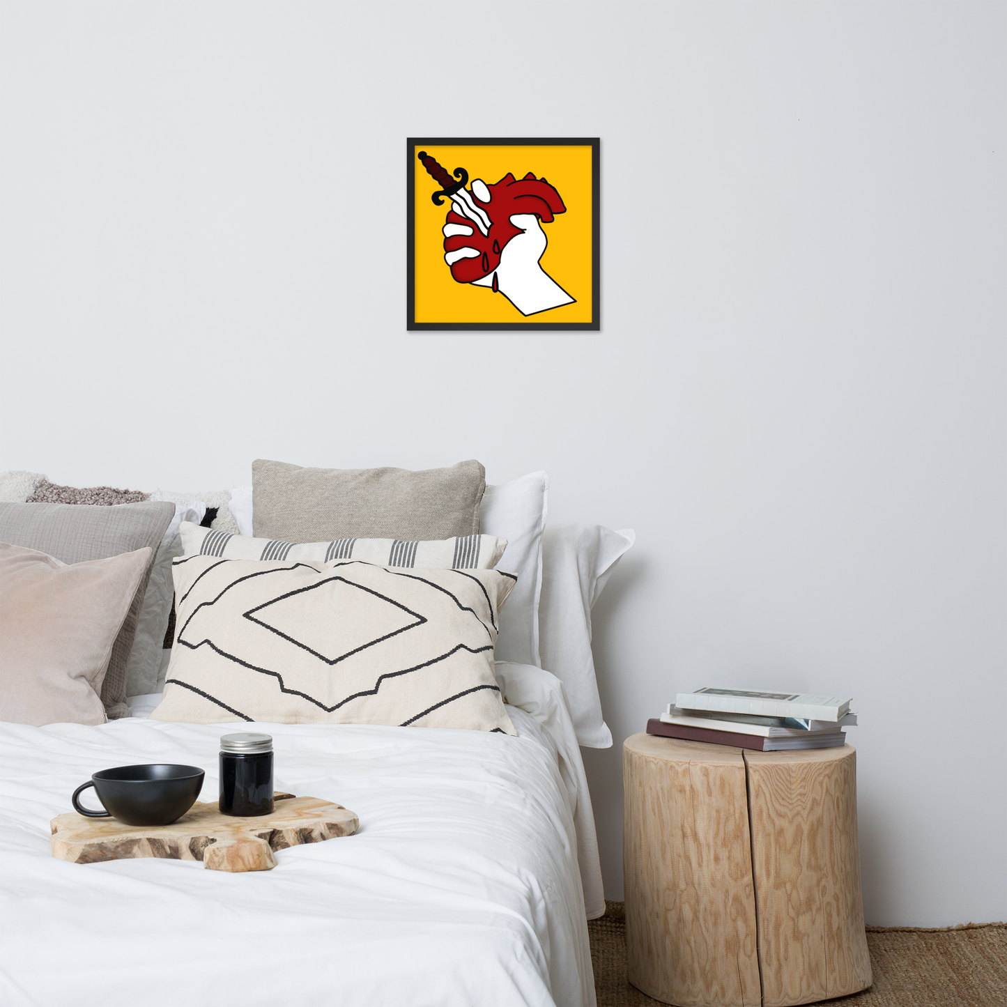 Bedroom with a bed, pillows, and a small table with a coffee cup and books, featuring a red, white, and yellow framed poster of a hand holding up a heart stabbed by a dagger on the wall.