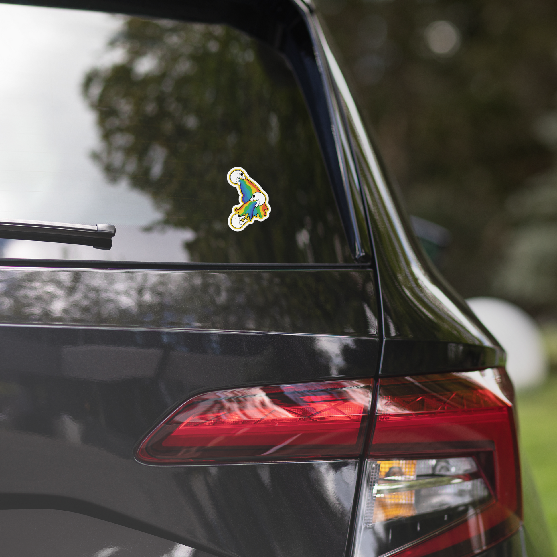 Colorful rainbow sticker of 3 skulls vomiting on the rear window of a black car with blurred greenery in the background