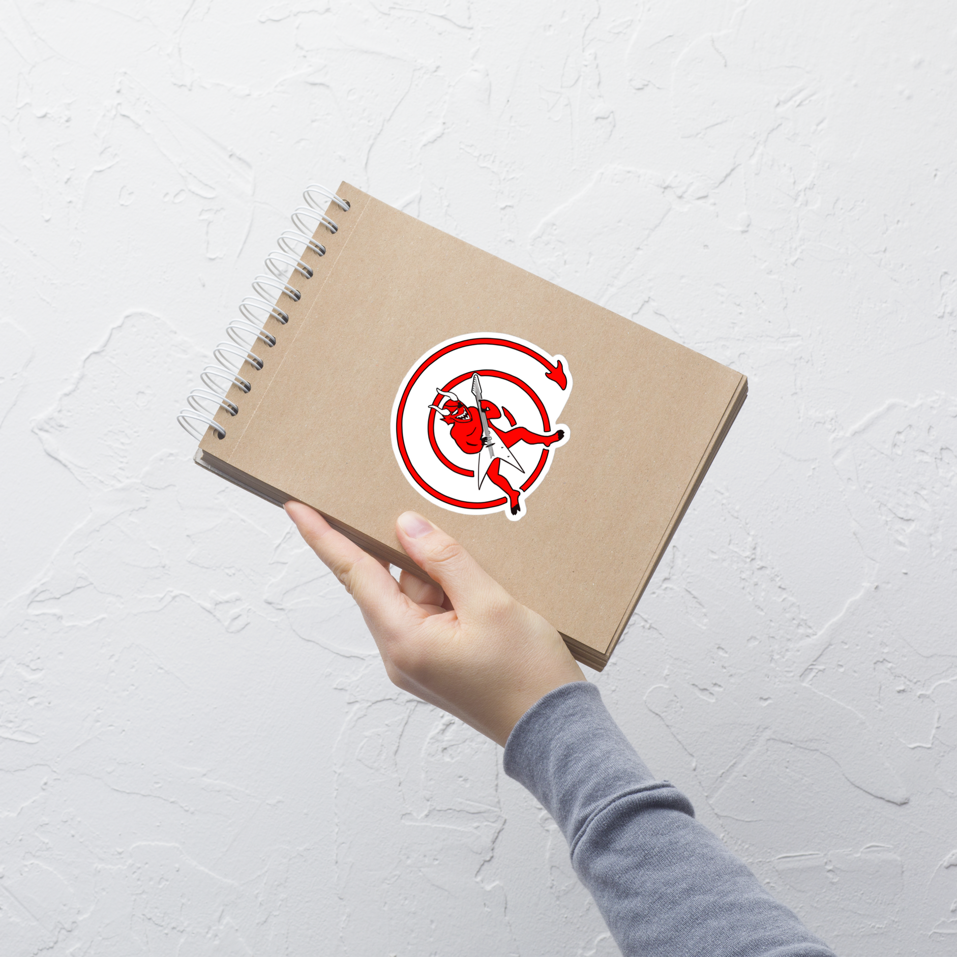 Hand holding a brown notebook with a sticker of a red devil with a guitar inside a red circle on a white background a light gray textured background