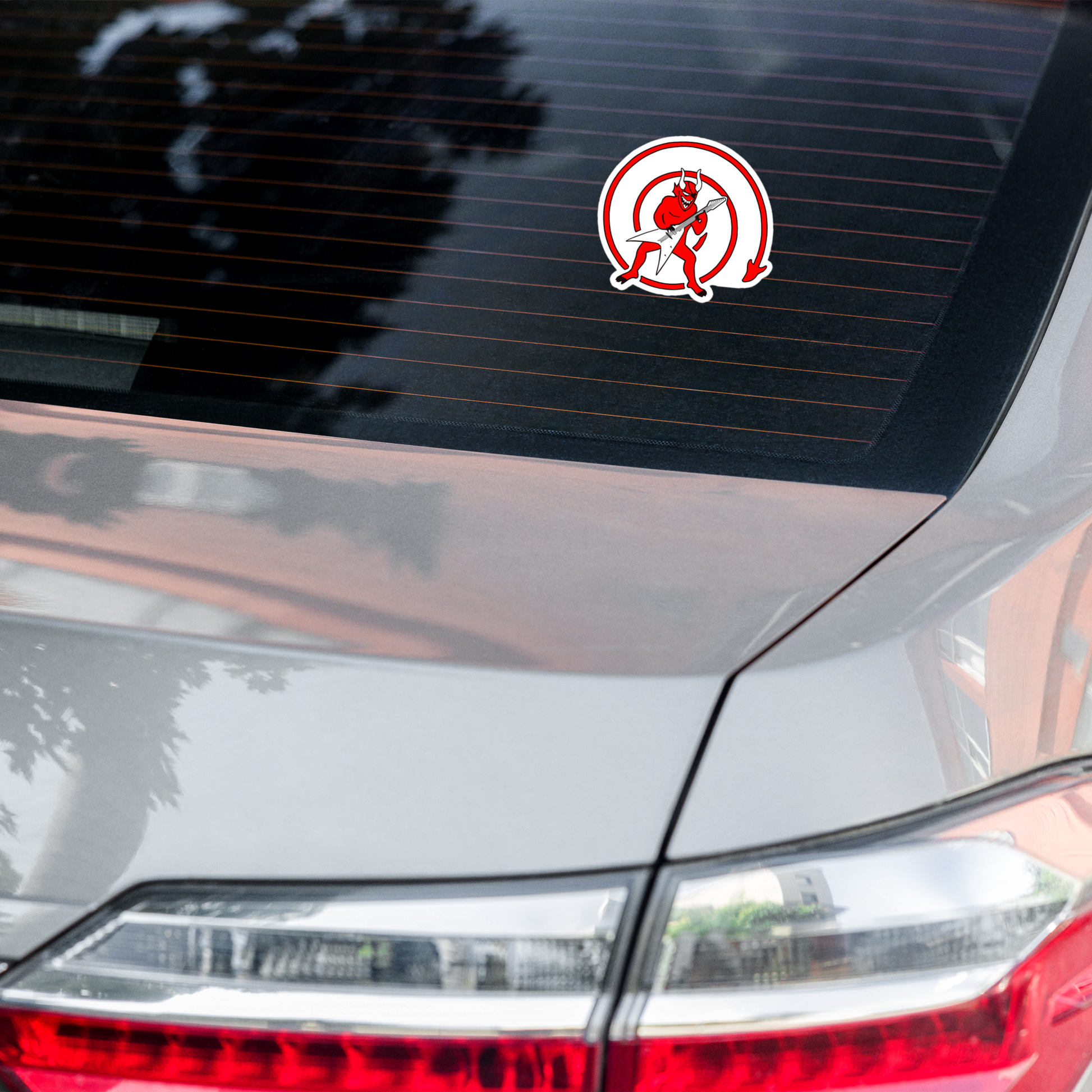 Car with a sticker of a red devil with a guitar inside a red circle on a white background on the rear window.