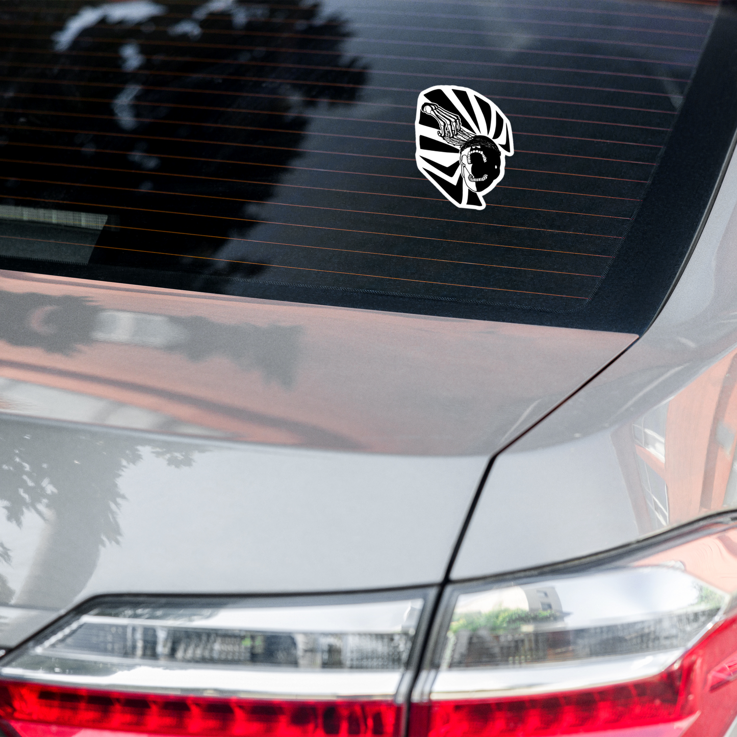 Car sticker with a black and white graphic of a skeleton hand reaching upwards to pull thorns from his face in front of a sharp pattern in the background on the rear window of a car