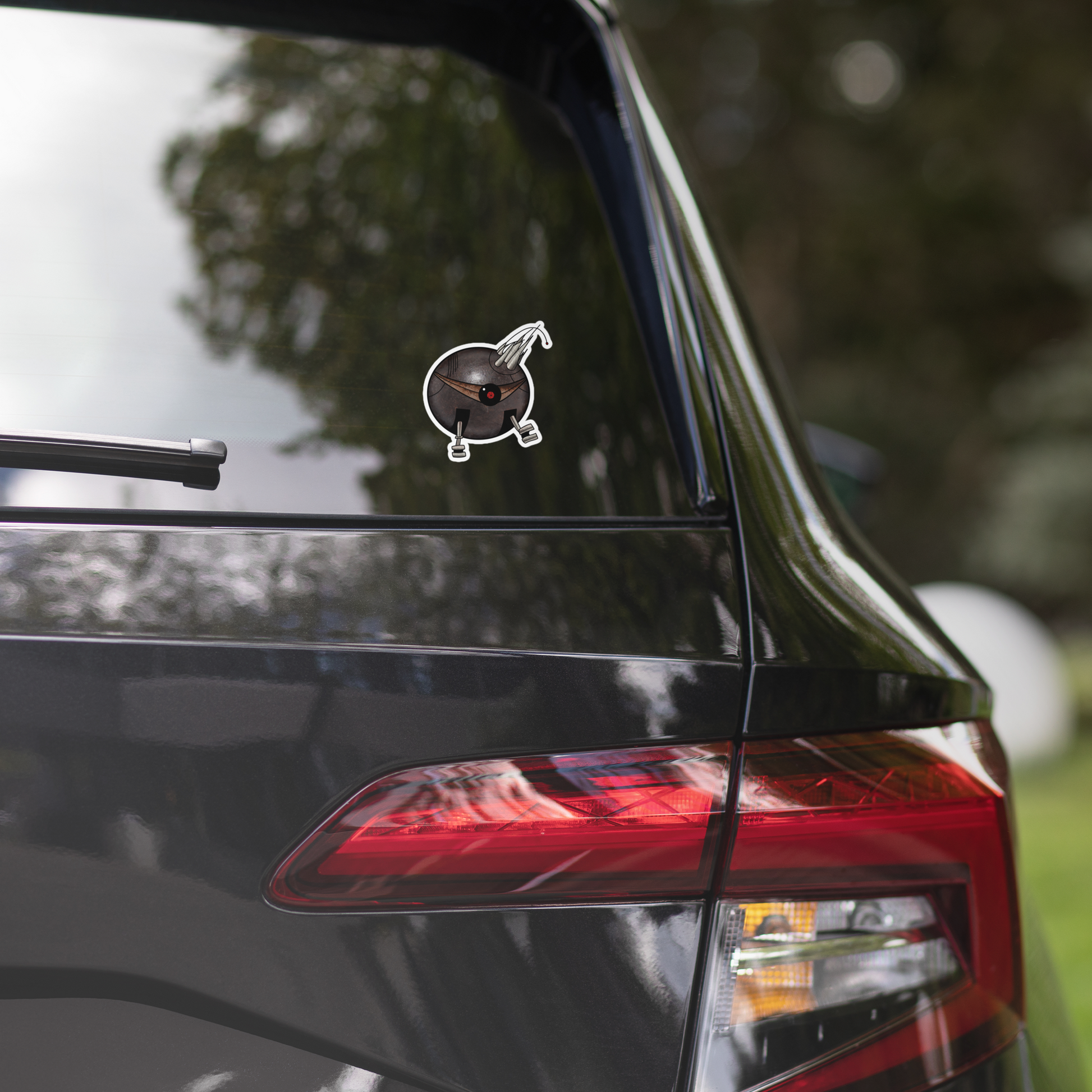 A die-cut vinyl sticker featuring a stylized, cartoonish round robot with a dark metallic gray body, a single large red and black eye, metallic silver legs, and a pair of curved metallic claws on its head, isolated on a white background on the back of a black SUV