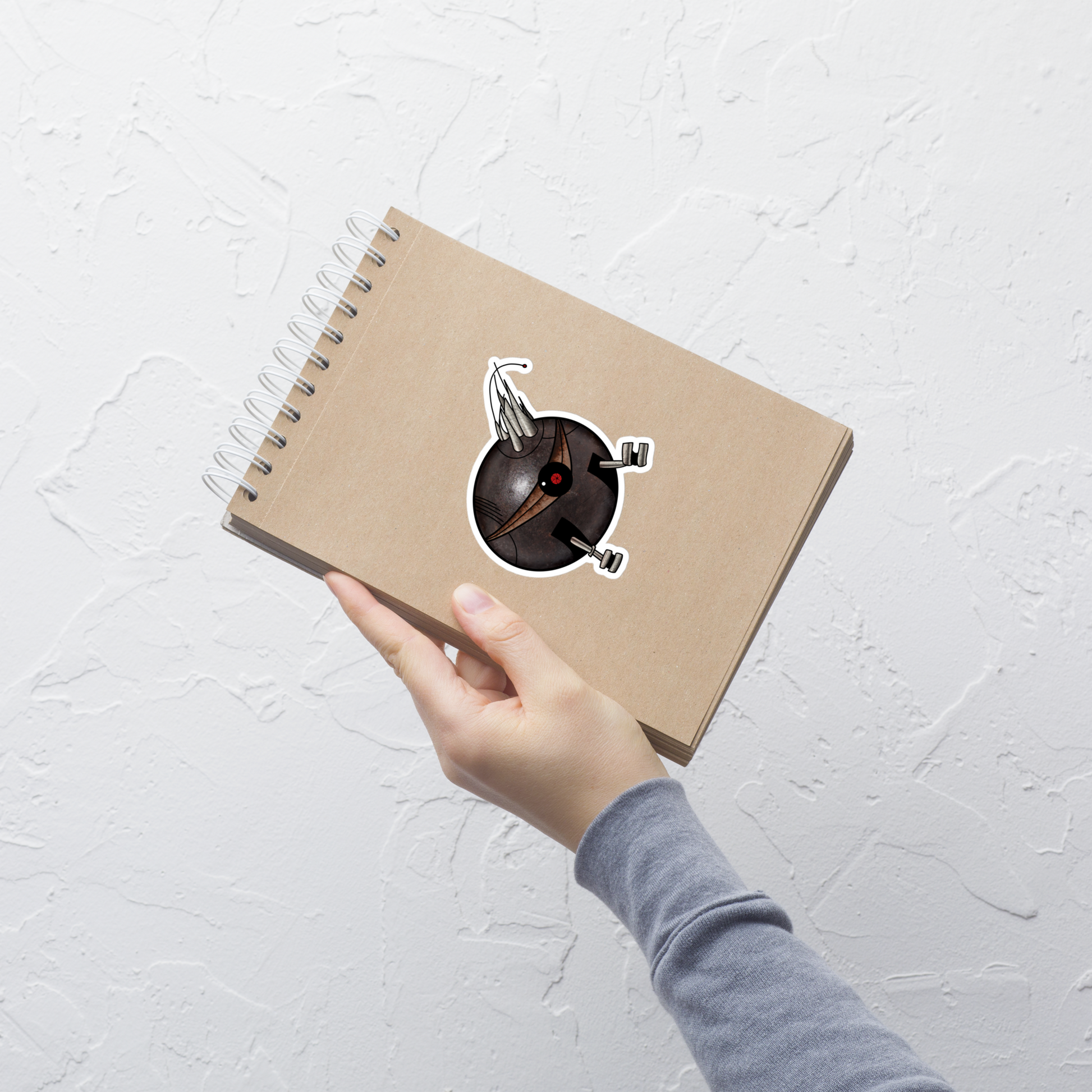 A hand holding a brown kraft paper spiral notebook against a textured white wall. The notebook features a die-cut sticker of a black spherical robot with a red center