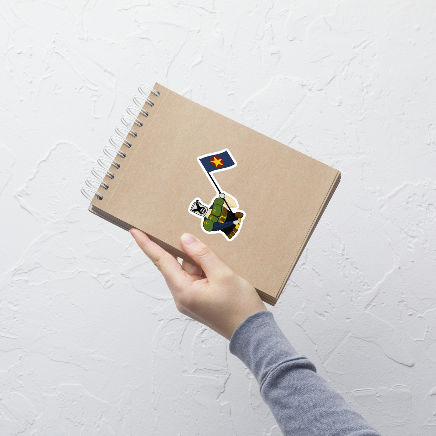 A hand holding a brown kraft paper spiral notebook against a textured white wall. The notebook has a die-cut sticker of a cartoon soldier holding a blue flag with a yellow star.