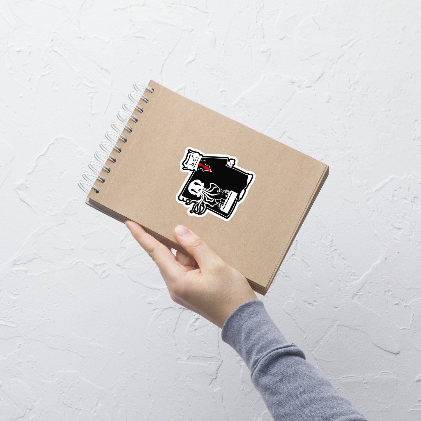 A hand wearing a gray long-sleeve shirt holds a spiral-bound notebook with a kraft paper cover. The notebook has a die-cut sticker on the front featuring a black and white illustration of a robot character in a trench coat