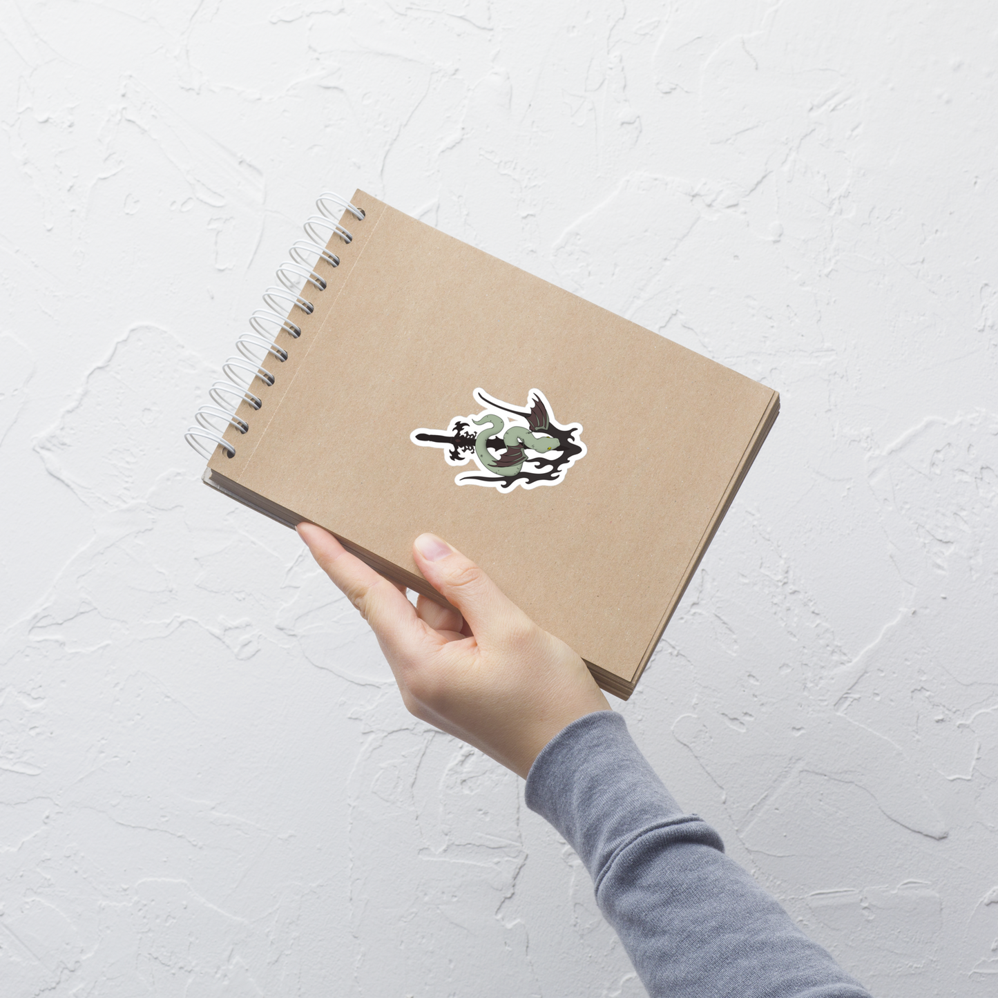 Hand holding a brown notebook with a die-cut vinyl sticker featuring a stylized green snake wrapped around a black sword, shown on a plain white background on a light gray textured background
