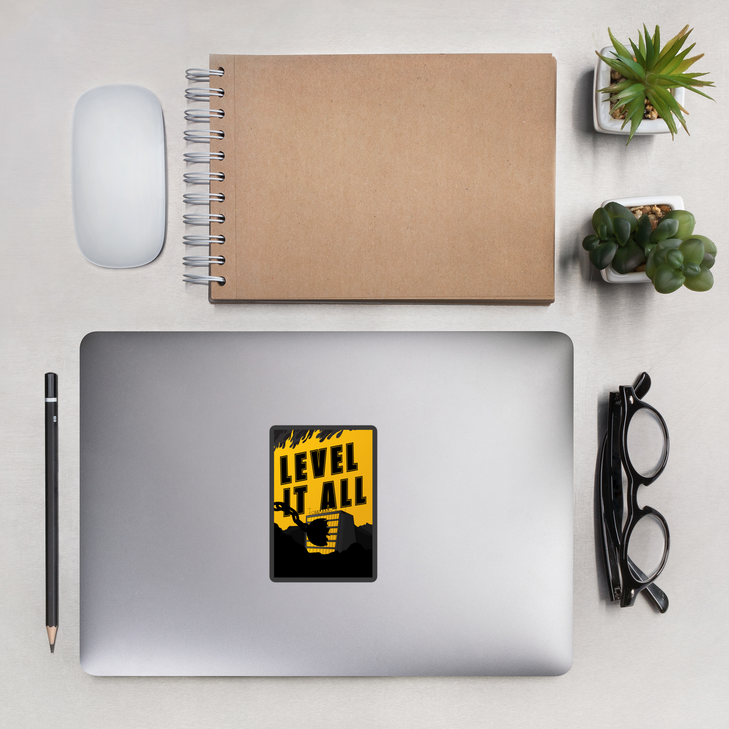 Laptop with a 'Level It All' sticker on a desk with a notebook, mouse, and plants.