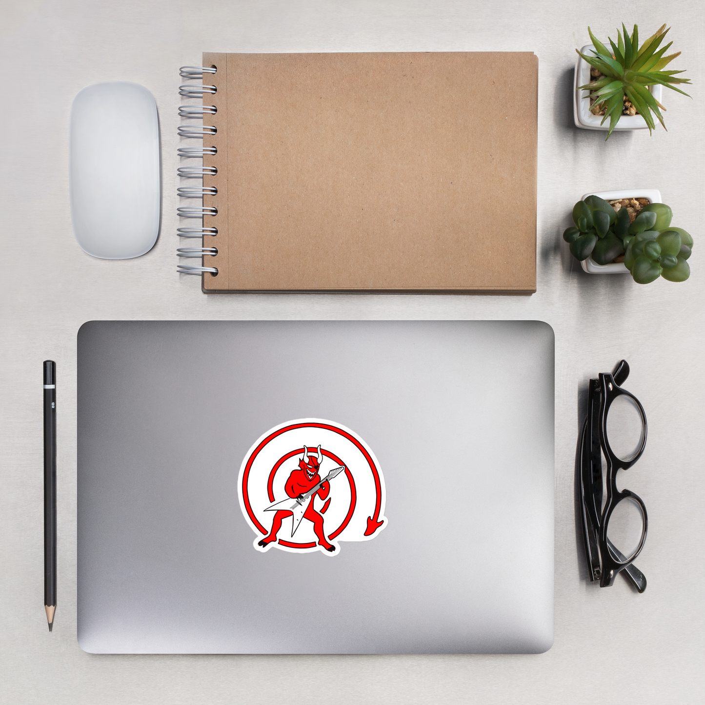 Laptop with a sticker of a red devil with a guitar inside a red circle on a white background on a desk with a notebook, mouse, and plants.