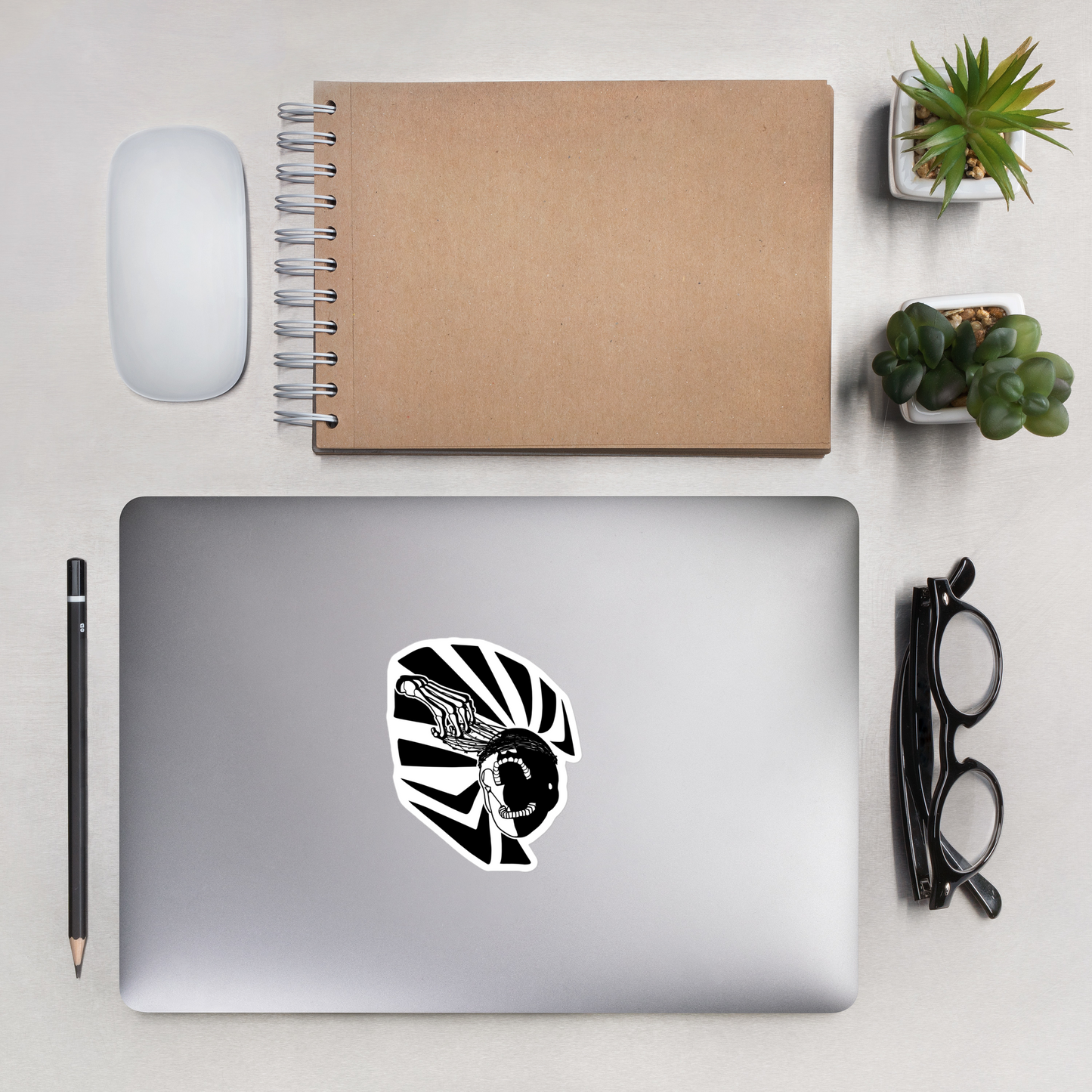 Laptop with a black and white graphic of a skeleton hand reaching upwards to pull thorns from his face in front of a sharp pattern in the background design on a desk with a notebook, mouse, and plants.