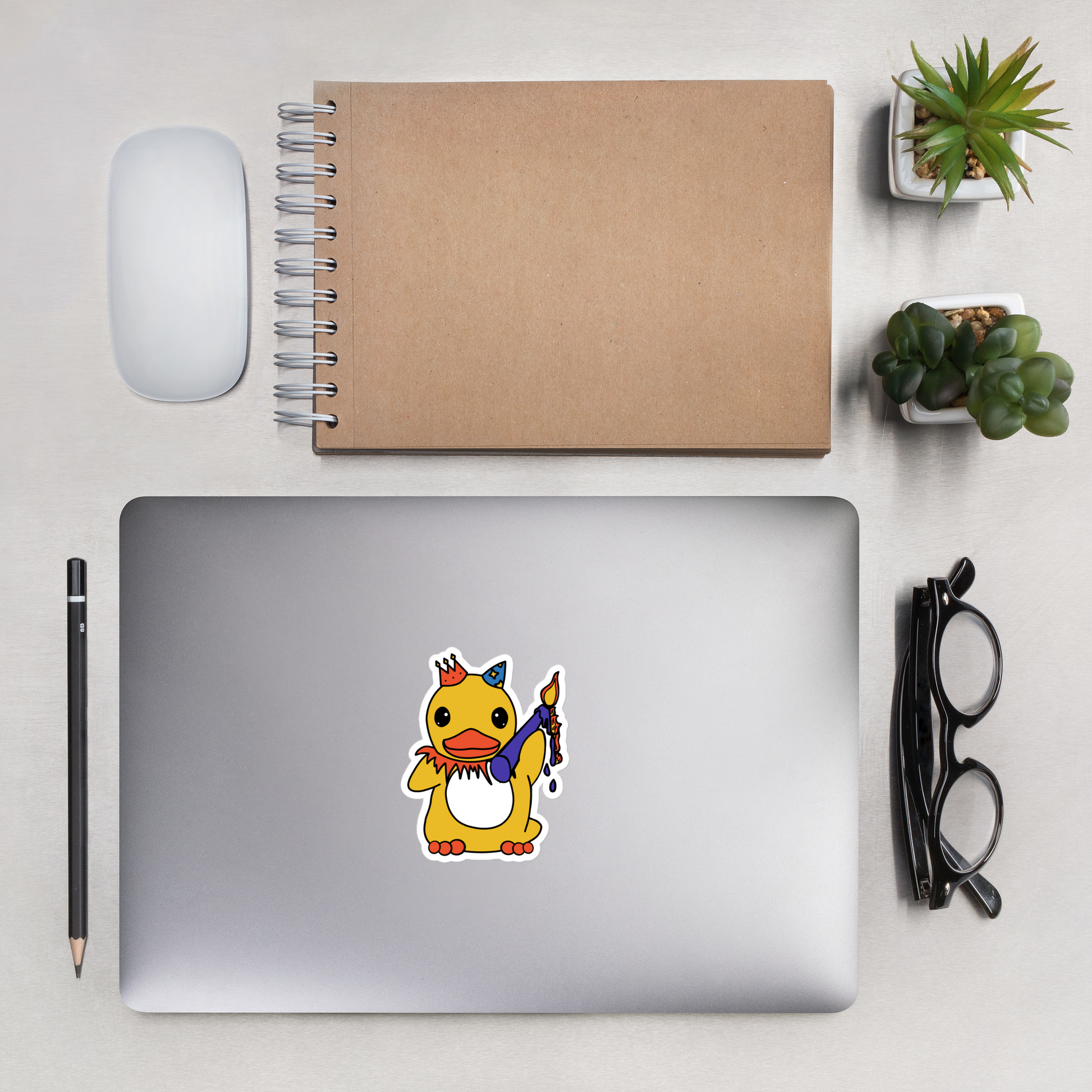 Laptop with a cartoon duck sticker on a desk with a notebook, mouse, pencil, and glasses.