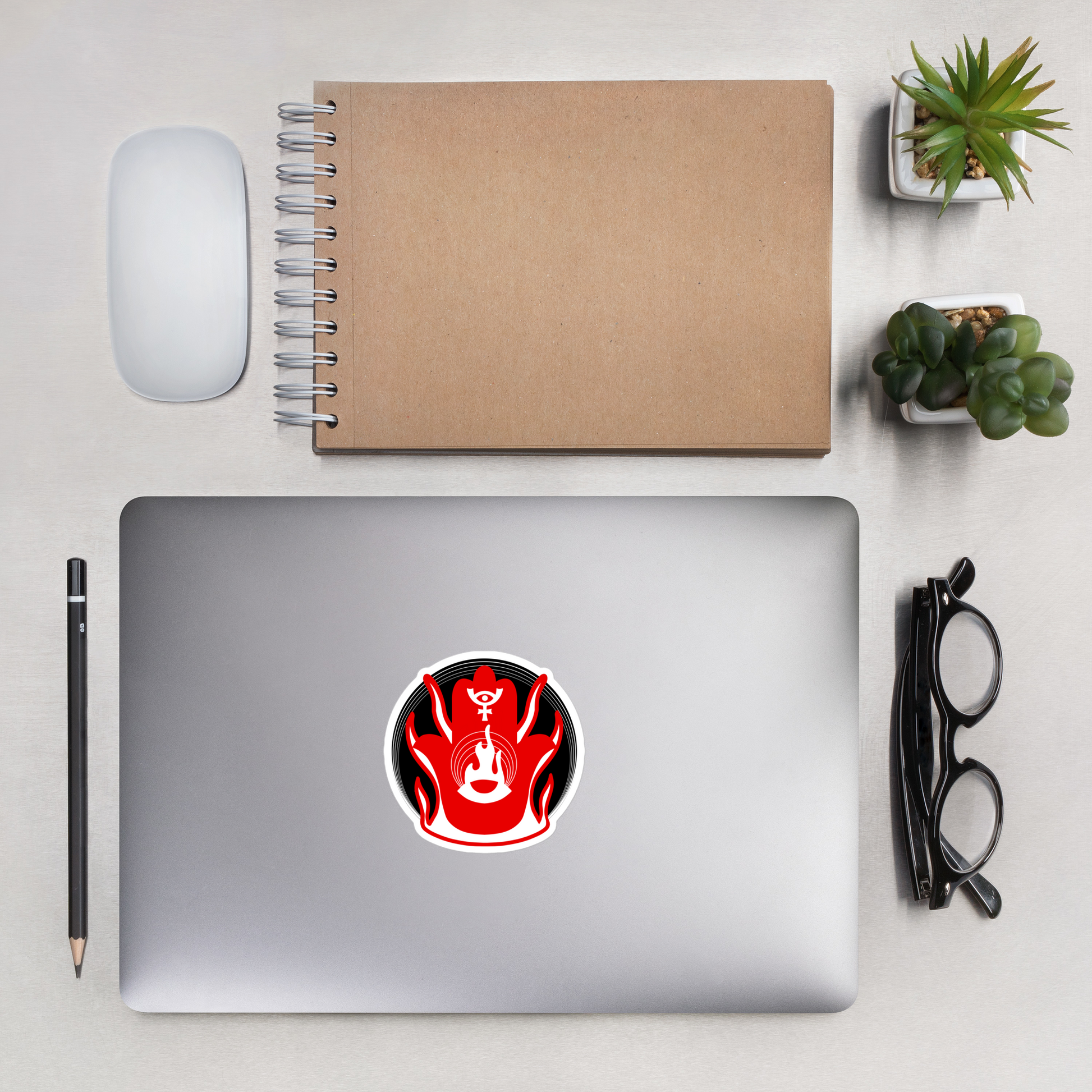Laptop with a red and black sticker on a desk with a notebook, mouse, pencil, and glasses.