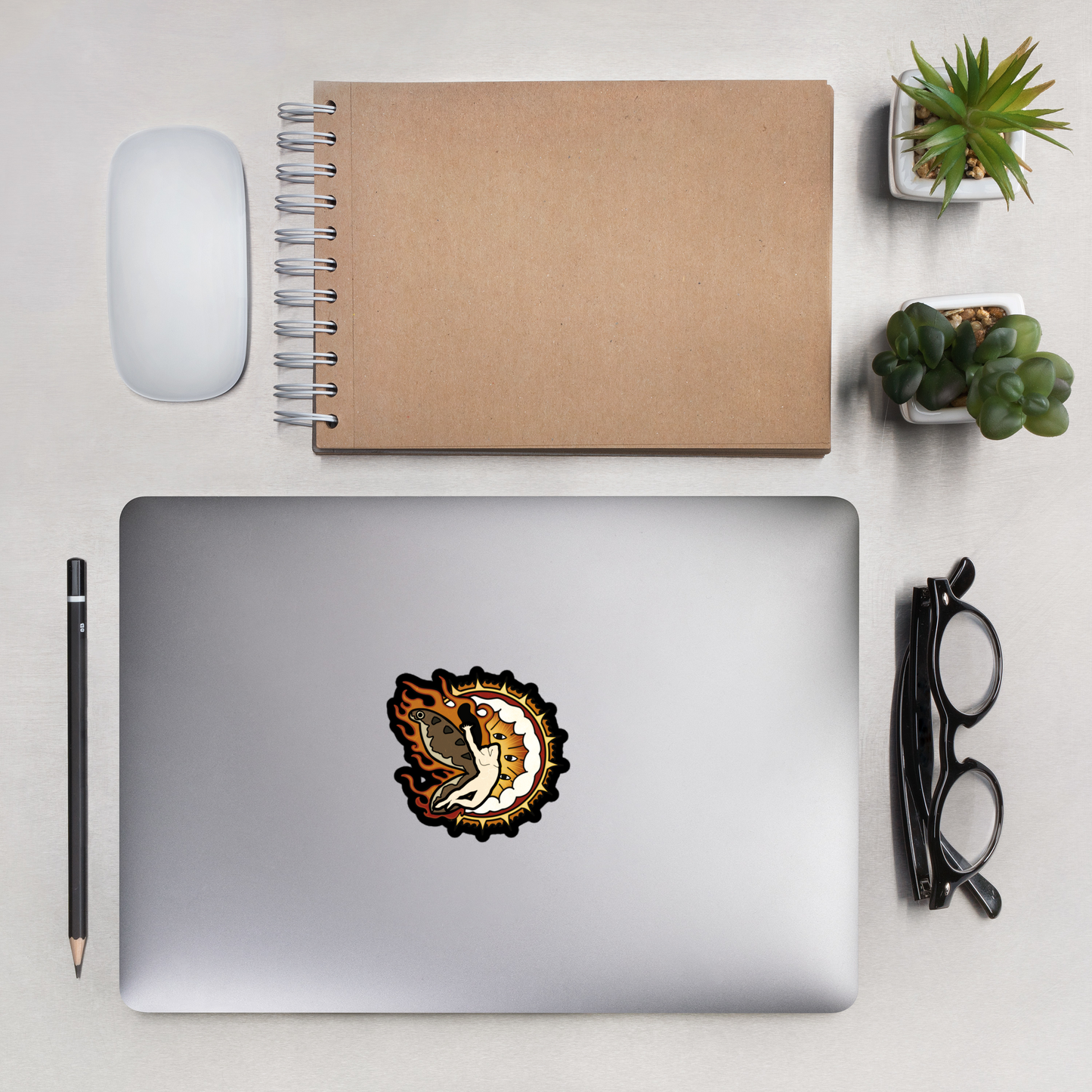 Sticker of a man with moth wings flying in front of a heavy metal biblical catholic design next to a journal, glasses, a pencil, two small succulents, and a computer mouse