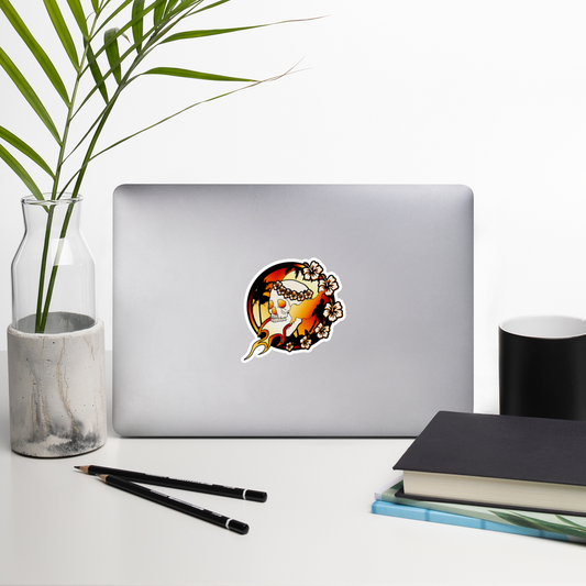 Laptop with a sticker of a skull with floral wreath and flames on a white background on a desk with a plant, pencils, and books.