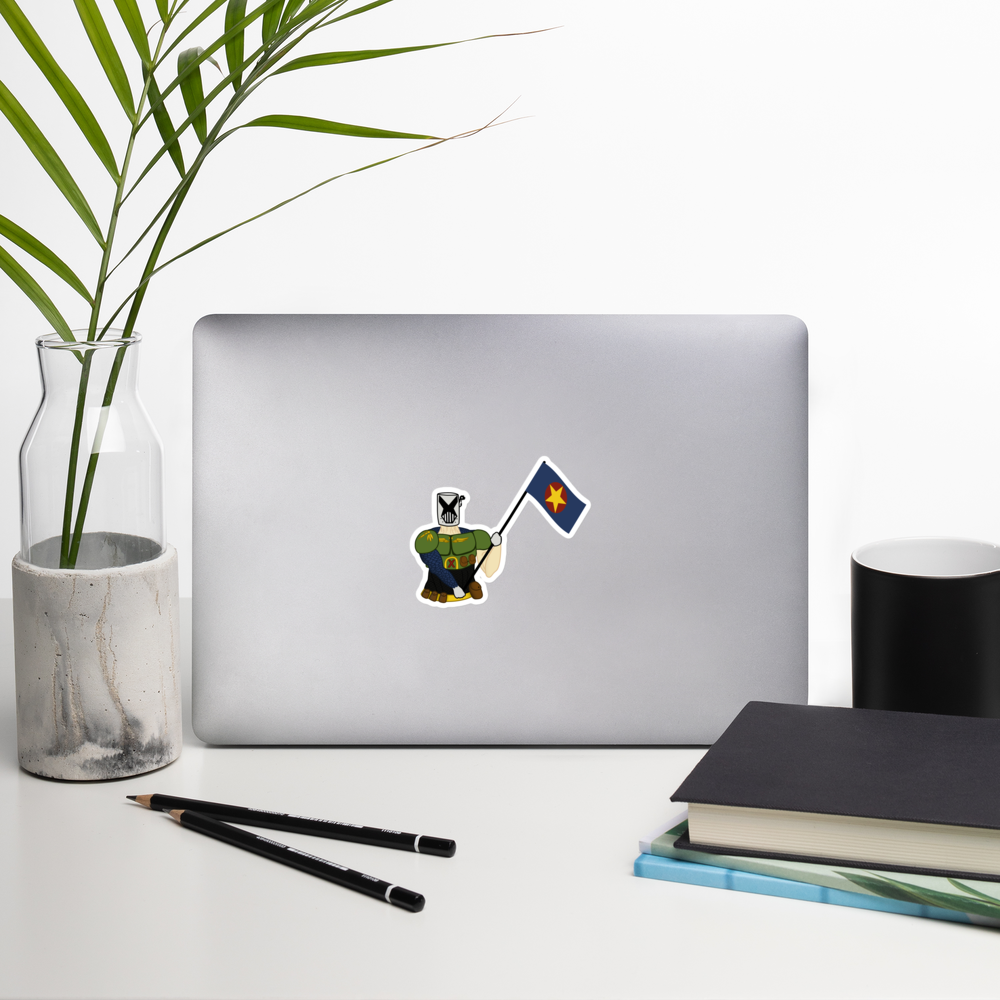 A silver laptop on a white desk with a small die-cut sticker of a cartoon soldier holding a blue flag with a yellow star. The scene is styled with a glass vase containing green palm leaves, two black pencils, stacked books, and a black cup.