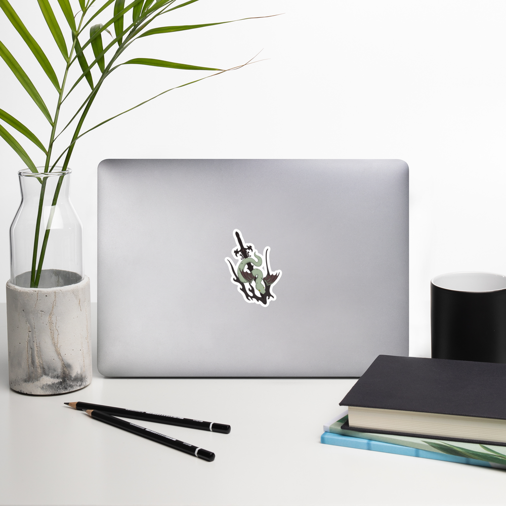 Laptop with a die-cut vinyl sticker featuring a stylized green snake wrapped around a black sword, shown on a plain white background on a desk with a plant, mug, and books.
