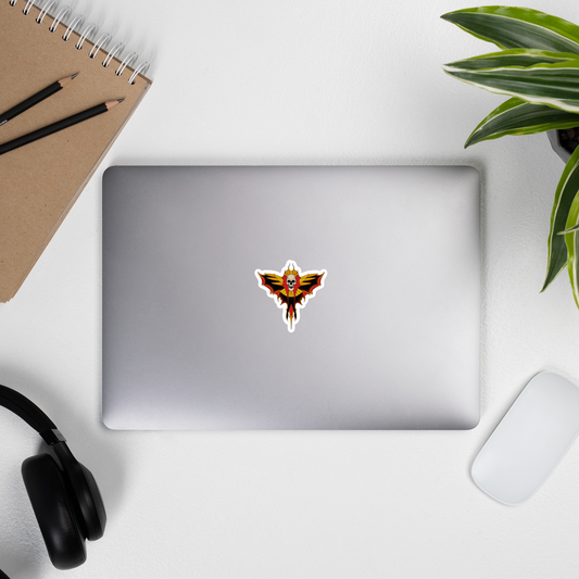 Laptop with a sticker with a tattoo skull moth design featuring wings on a white background on a desk with a notebook, pens, headphones, and a plant.