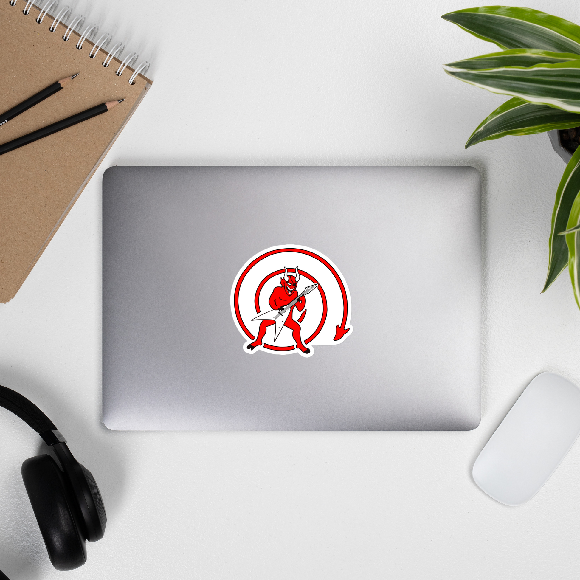 Laptop with a red and white sticker of a cartoonish demon playing the guitar on a desk with a notebook, pencils, headphones, and a plant.