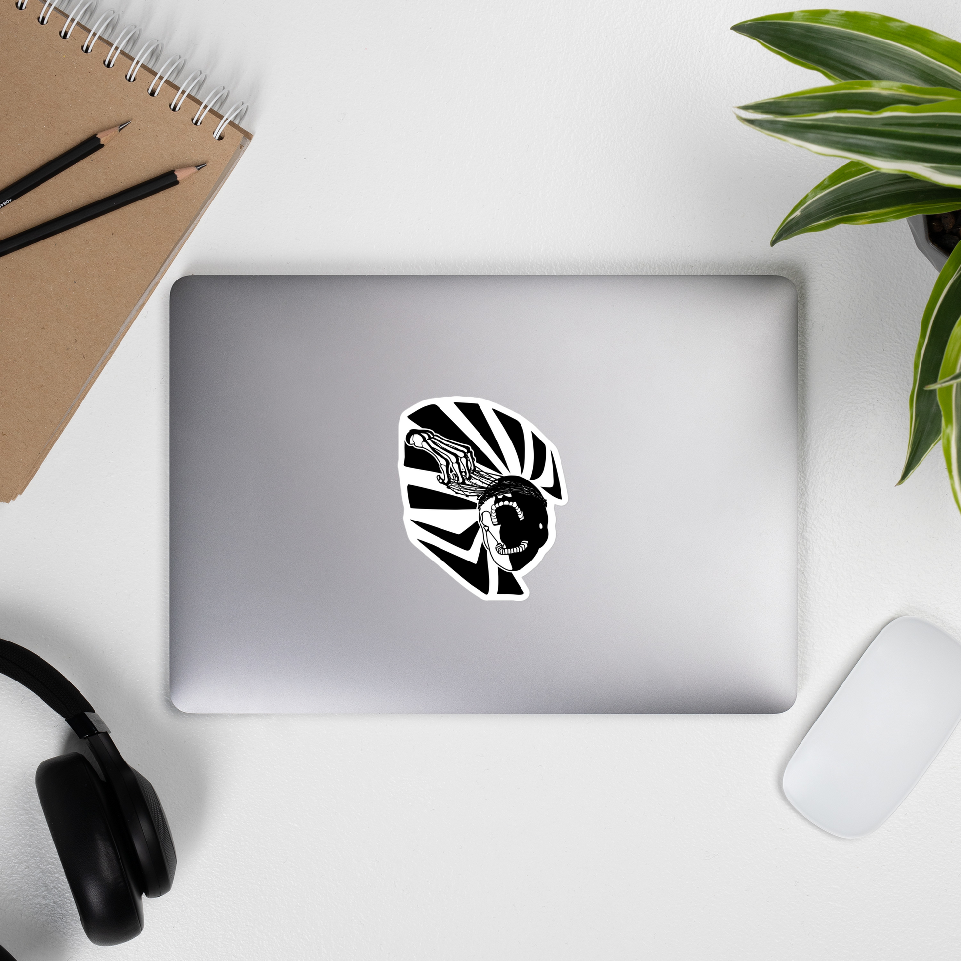 Laptop with a black and white graphic of a skeleton hand reaching upwards to pull thorns from his face in front of a sharp pattern in the background on a desk with a notebook, pencils, and headphones.
