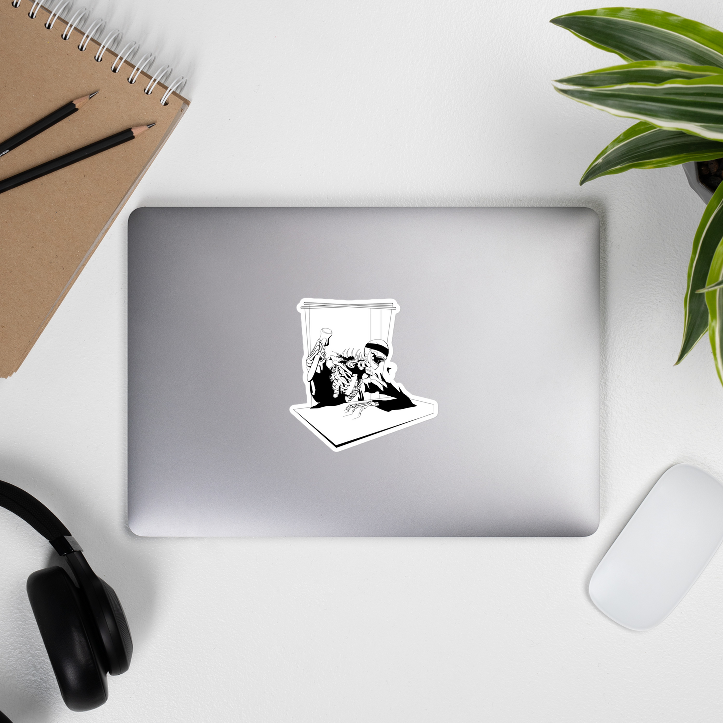 Laptop with a sticker of a black and white heavy metal style skeleton figure with a blindfold holding a gavel, sitting on a chair with a table in front on a desk with a notebook, pencils, headphones, and a plant.