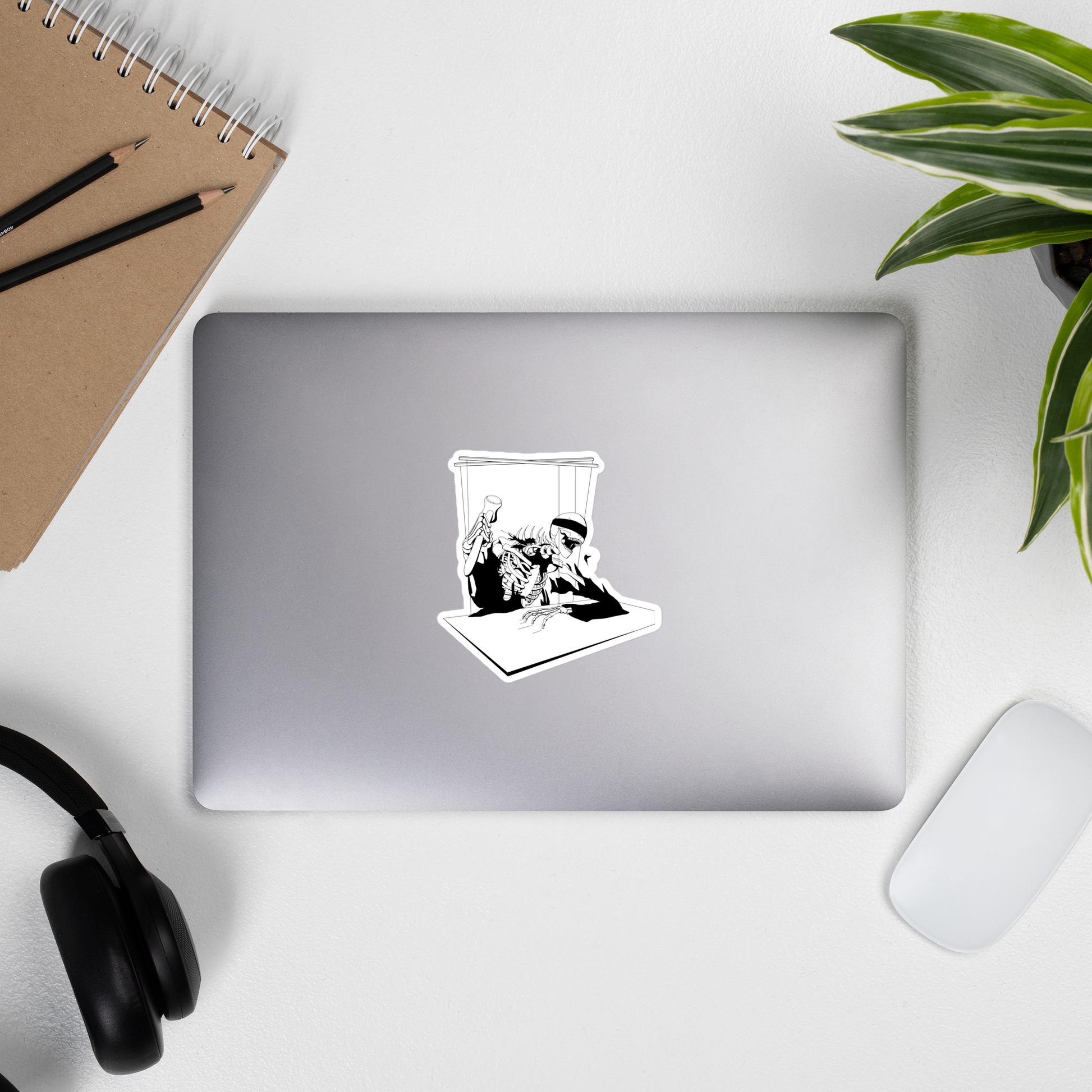 Laptop with a sticker of a black and white heavy metal style skeleton figure with a blindfold holding a gavel, sitting on a chair with a table in front on a desk with a notebook, pencils, headphones, and a plant.