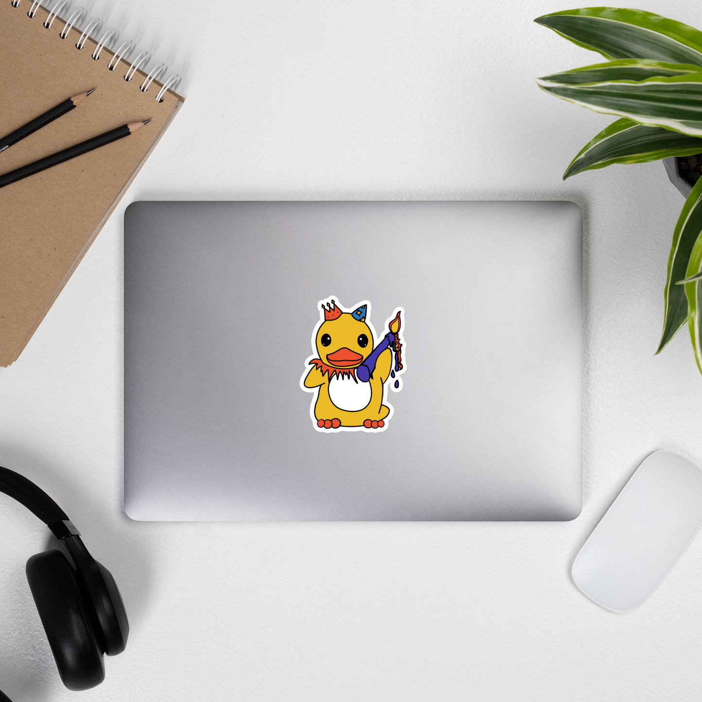 Laptop with a cartoon duck sticker on a desk with a notebook, pencils, and headphones.