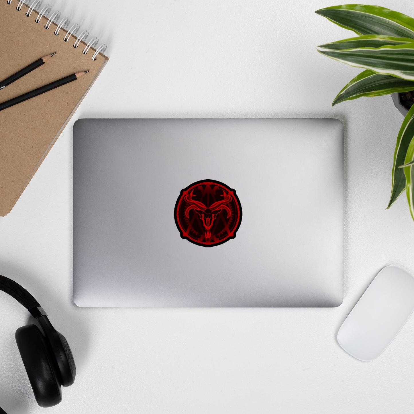 Laptop with a red and black logo on a white surface with a notebook, pens, and plant.