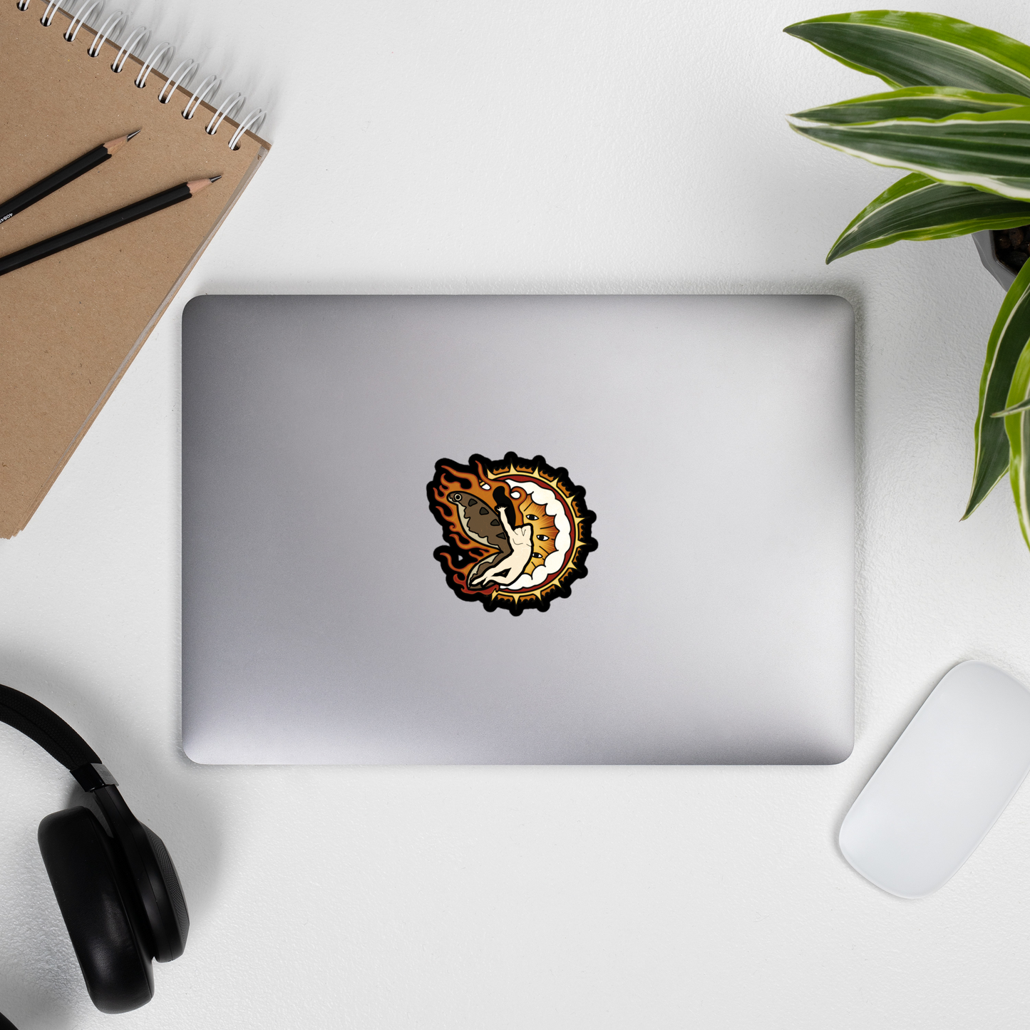 Sticker of a man with moth wings flying in front of a heavy metal biblical catholic design on a laptop next to some headphones, a journal with pencils on it, and a plant