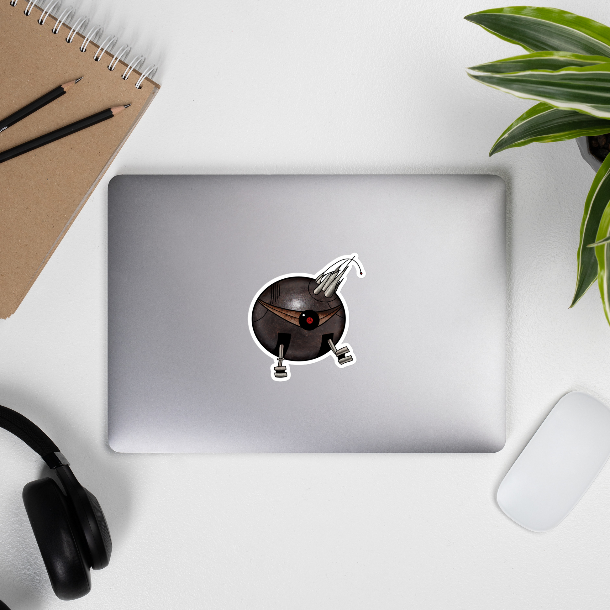 A die-cut vinyl sticker featuring a stylized, cartoonish round robot with a dark metallic gray body, a single large red and black eye, metallic silver legs, and a pair of curved metallic claws on its head, isolated on a white background on a laptop by a computer mouse, a potted plant, headphones, and a journal