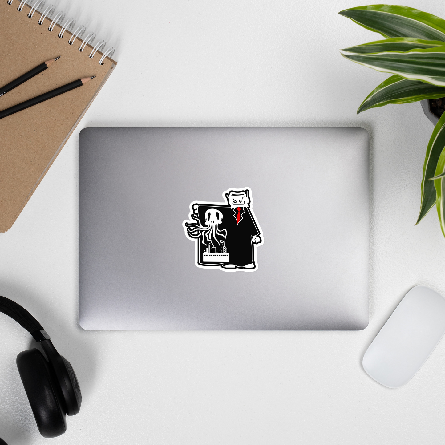 A die-cut vinyl sticker featuring a cartoon cat in a black suit holding a framed picture, shown applied to the back of a silver laptop. The laptop is part of a styled desk flat-lay with a notebook, headphones, a mouse, and a potted plant.