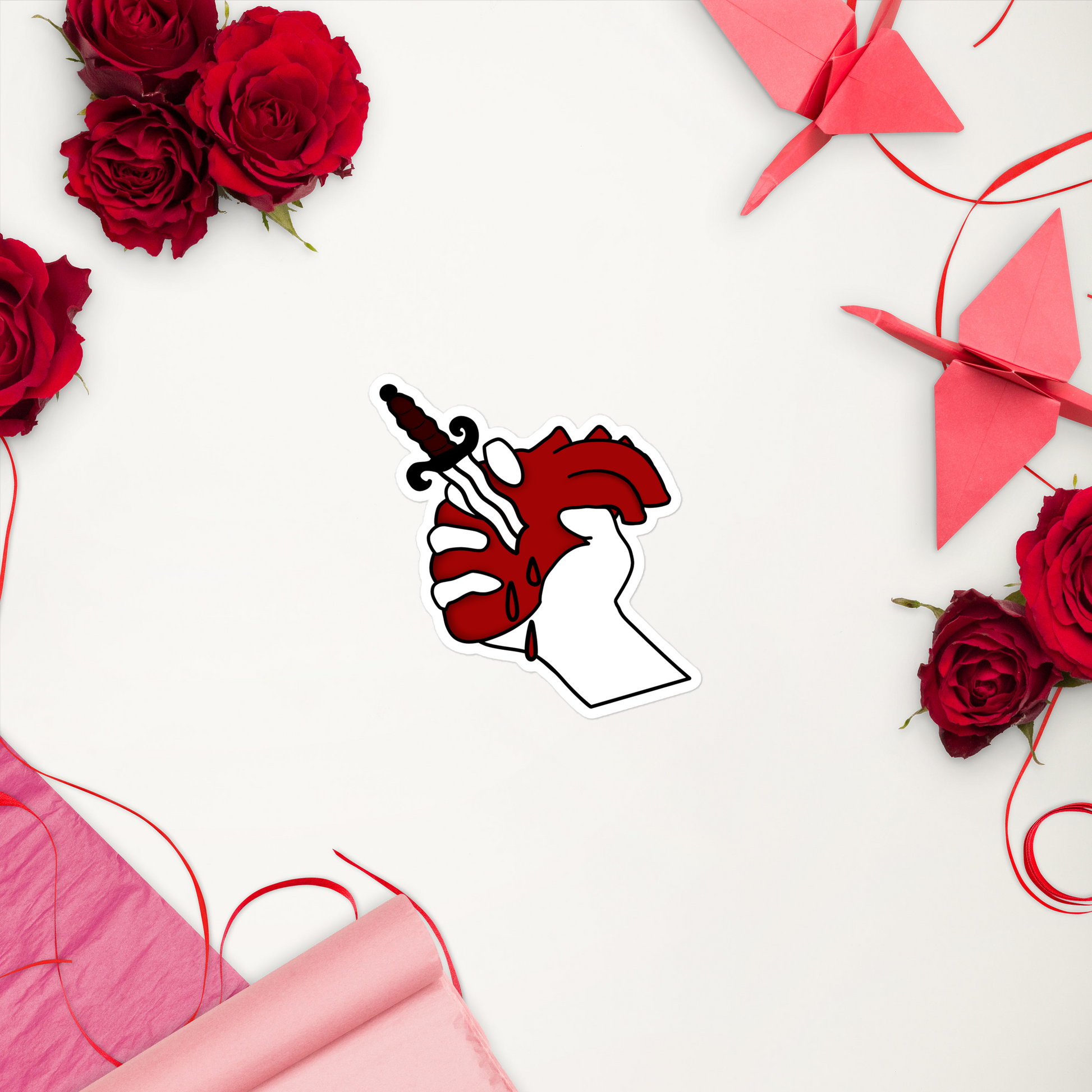 Decorative arrangement with red roses, pink ribbons, and a heart-shaped sticker on a white background