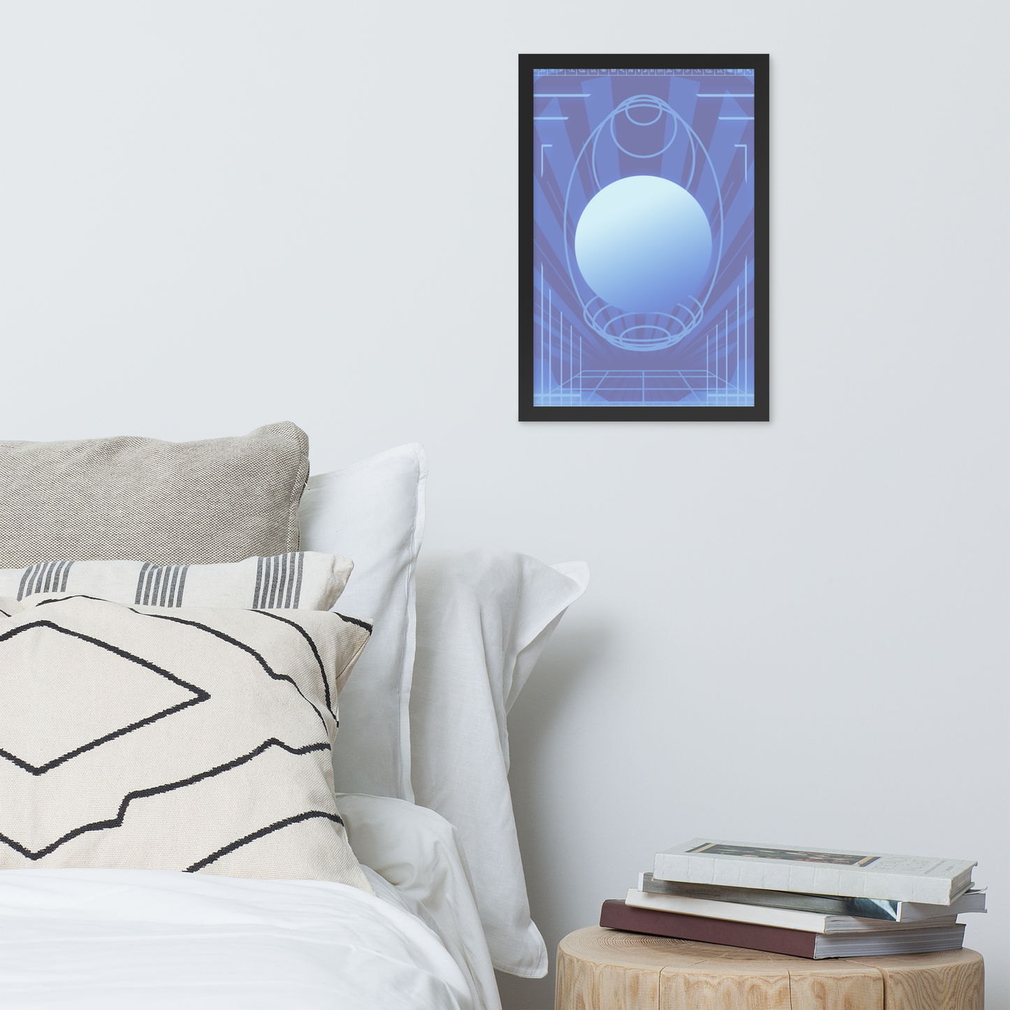 A framed abstract art print featuring a glowing blue sphere and geometric lines on a blue background, displayed on a light gray wall above a bed with pillows and a small wooden side table with books.