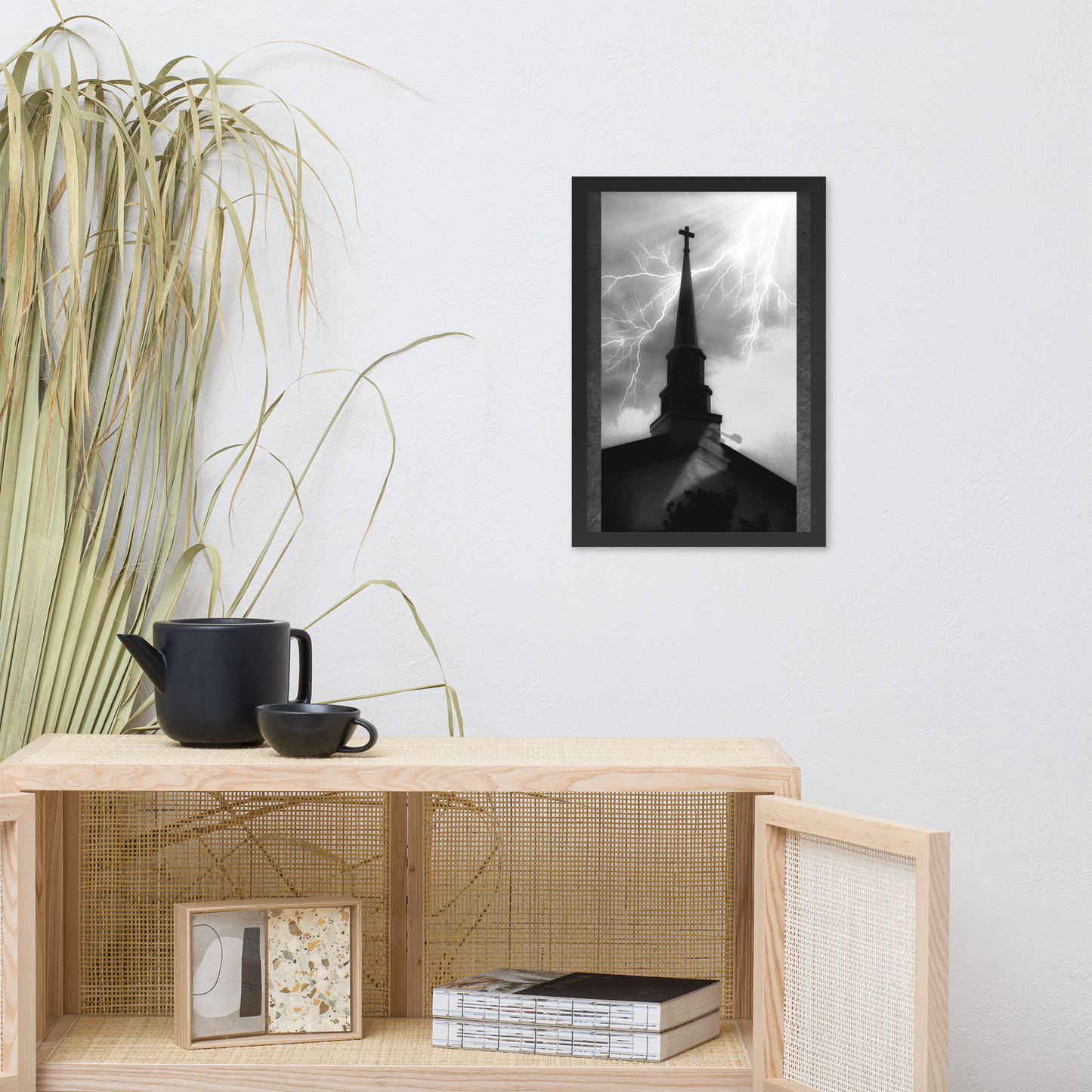 A black and white framed art print depicting a church steech silhouette under a stormy sky with lightning, displayed on a white wall above a light wood shelf with decorative items.