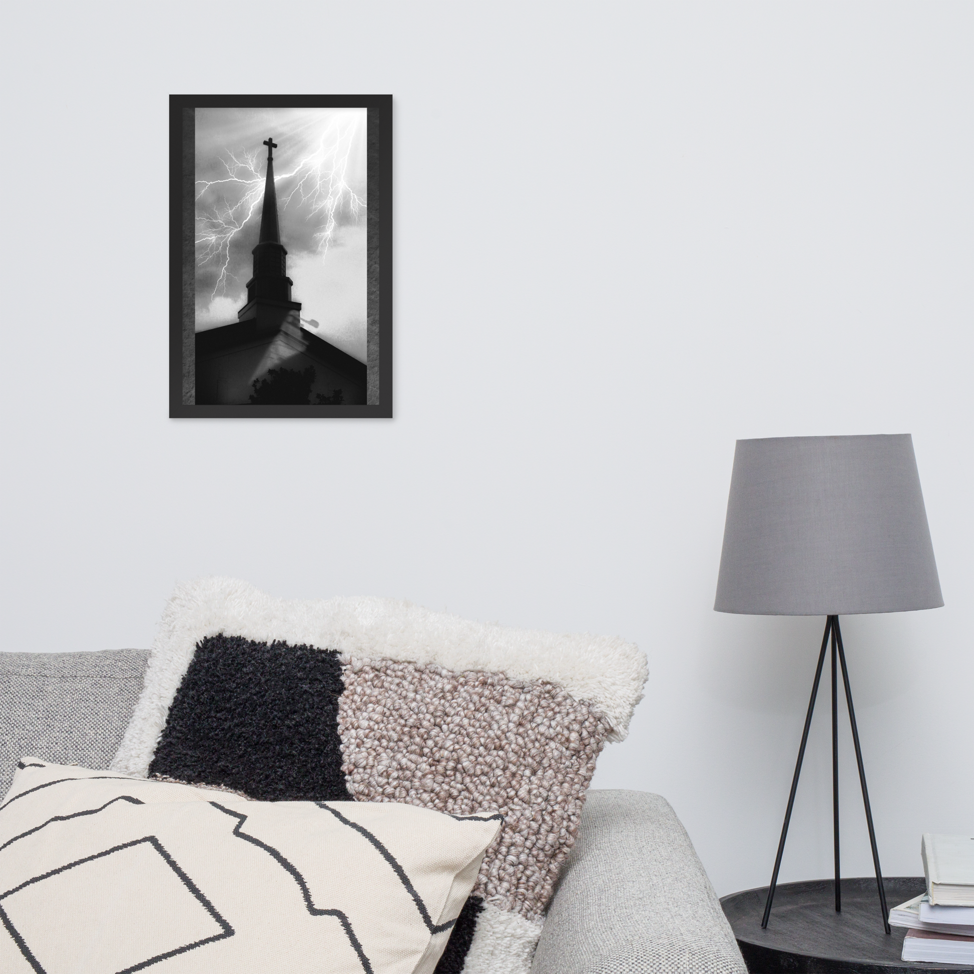 A framed black and white photograph of a church steeple with dramatic lightning, displayed on a light gray wall above a sofa with textured pillows and a floor lamp nearby.