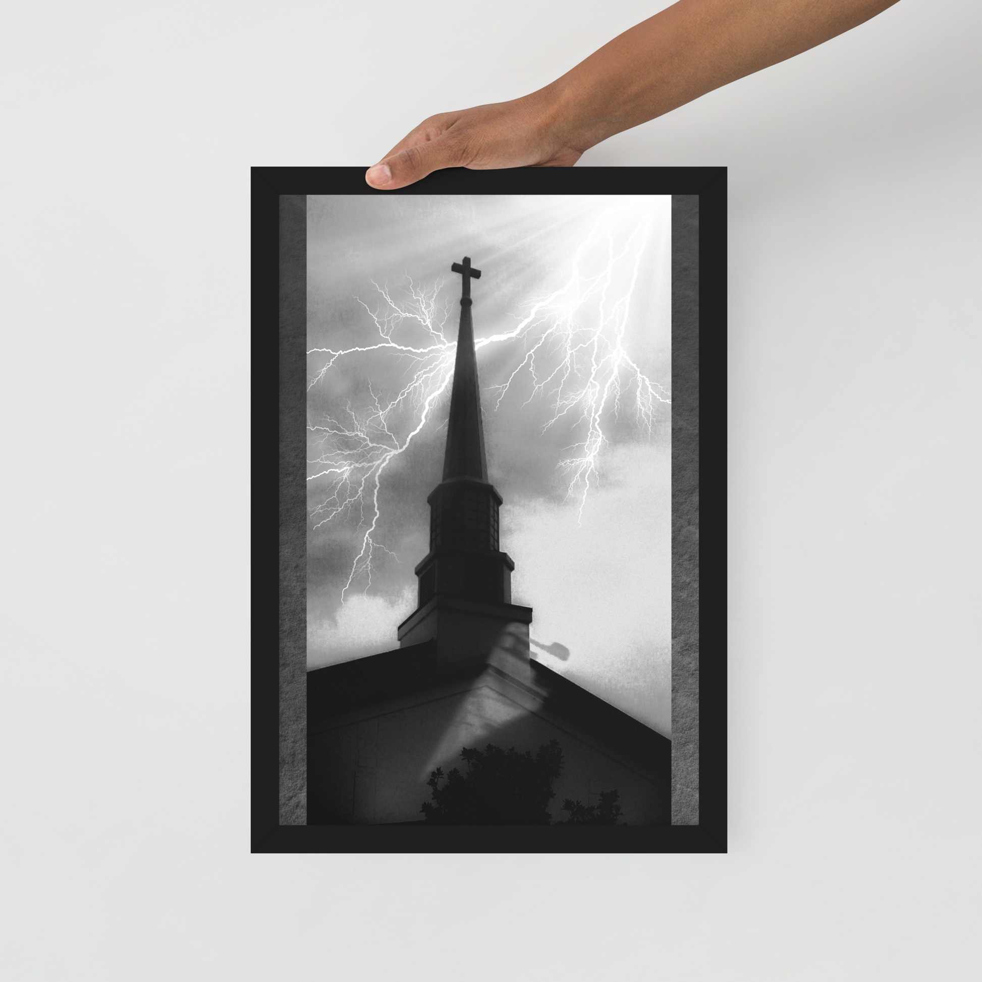 A framed black and white art print depicting a church steeple with lightning in the sky, held by a person's hand against a light gray wall.