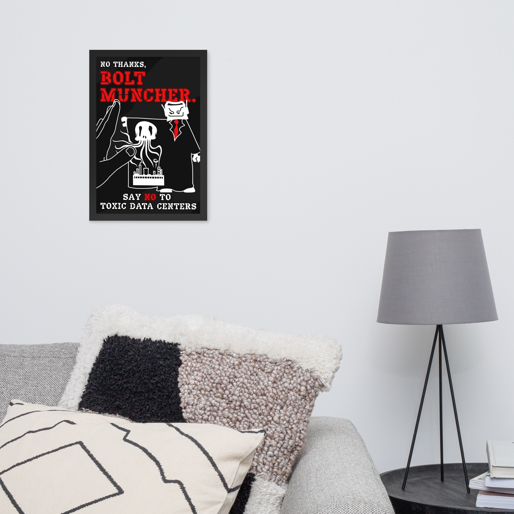 A black rectangular wall art print with red and white text and an illustration hangs on a light gray wall above a sofa with textured pillows. A gray floor lamp stands beside it in a styled interior setting.