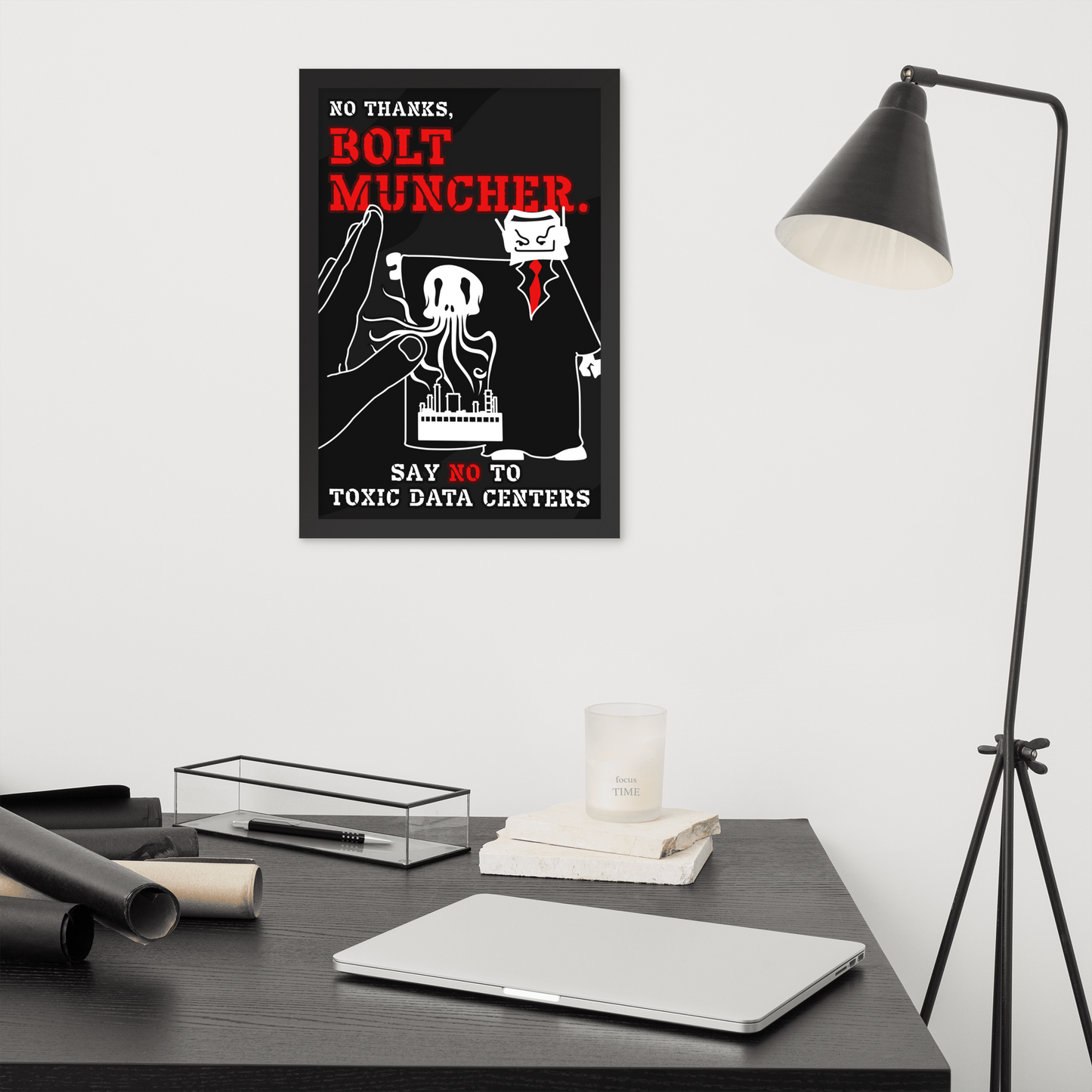 A framed black-and-white poster with bold red text reading 'BOLT MUNCHER' hangs on a light gray wall above a dark desk. The scene includes a black floor lamp, a closed silver laptop, rolled materials, a clear tray, and a small candle.