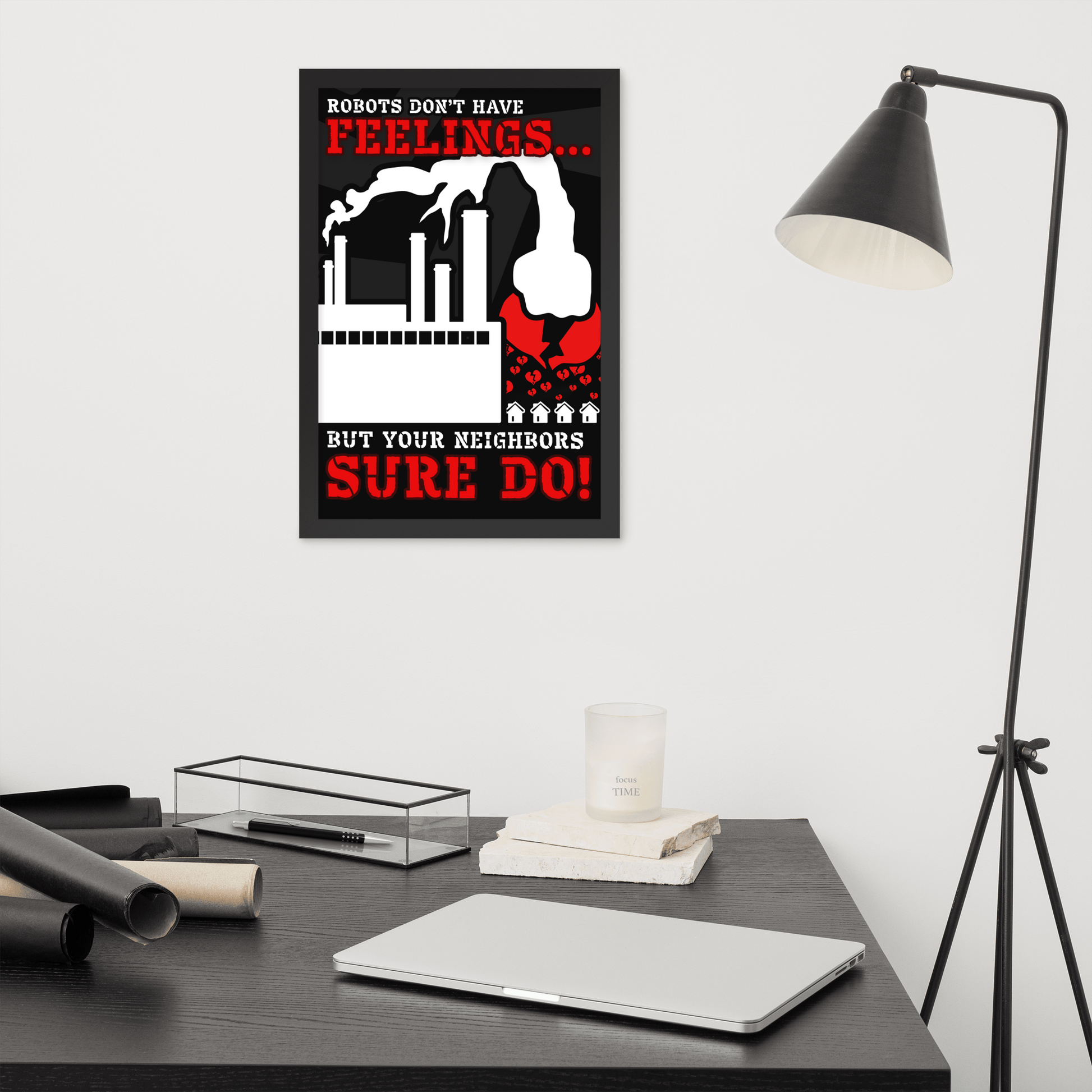 A framed wall art print with a black, white, and red robot-themed slogan hangs on a light gray wall above a dark desk with a laptop, lamp, and other office items.