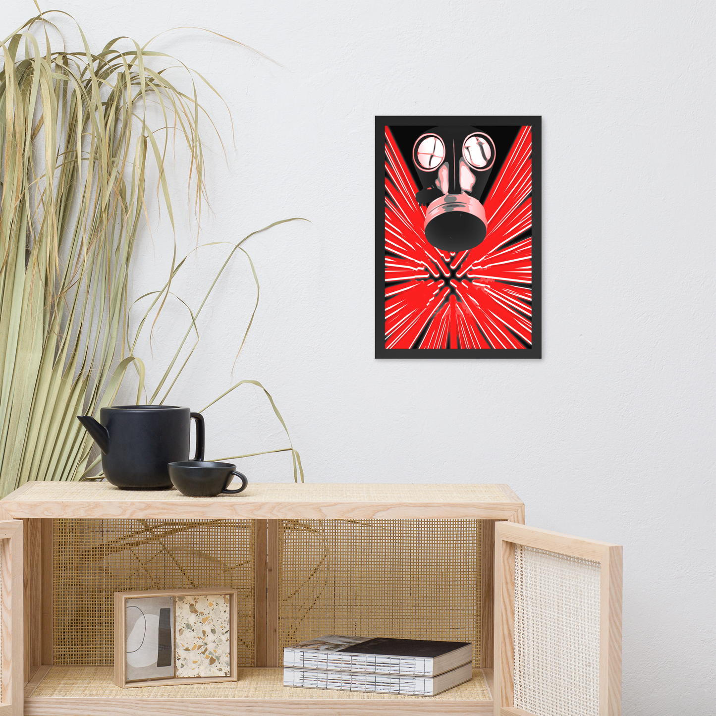 A framed wall art print featuring a stylized red and black gas mask with radiating red lines, displayed on a light gray wall above a wooden shelf with decorative items.
