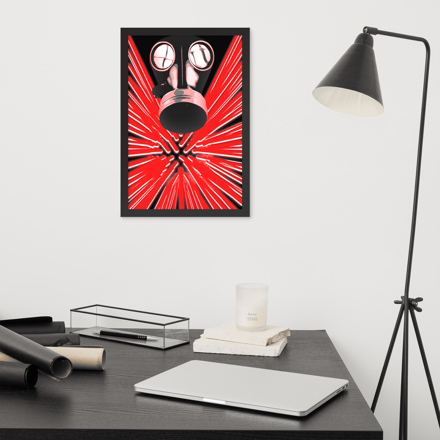 A framed wall art print featuring a stylized red and black gas mask with radiating lines, displayed above a dark desk in a modern room with a black floor lamp, laptop, and books.