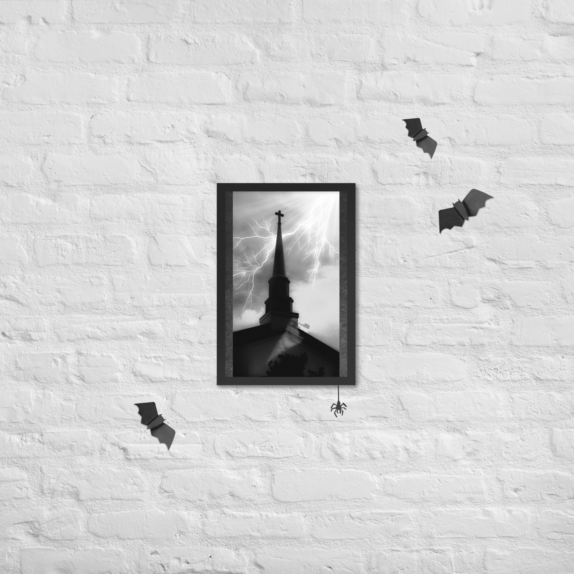 A black-framed black and white art print of a church steeple with lightning, displayed on a white brick wall with Halloween decorations including bats and a spider.