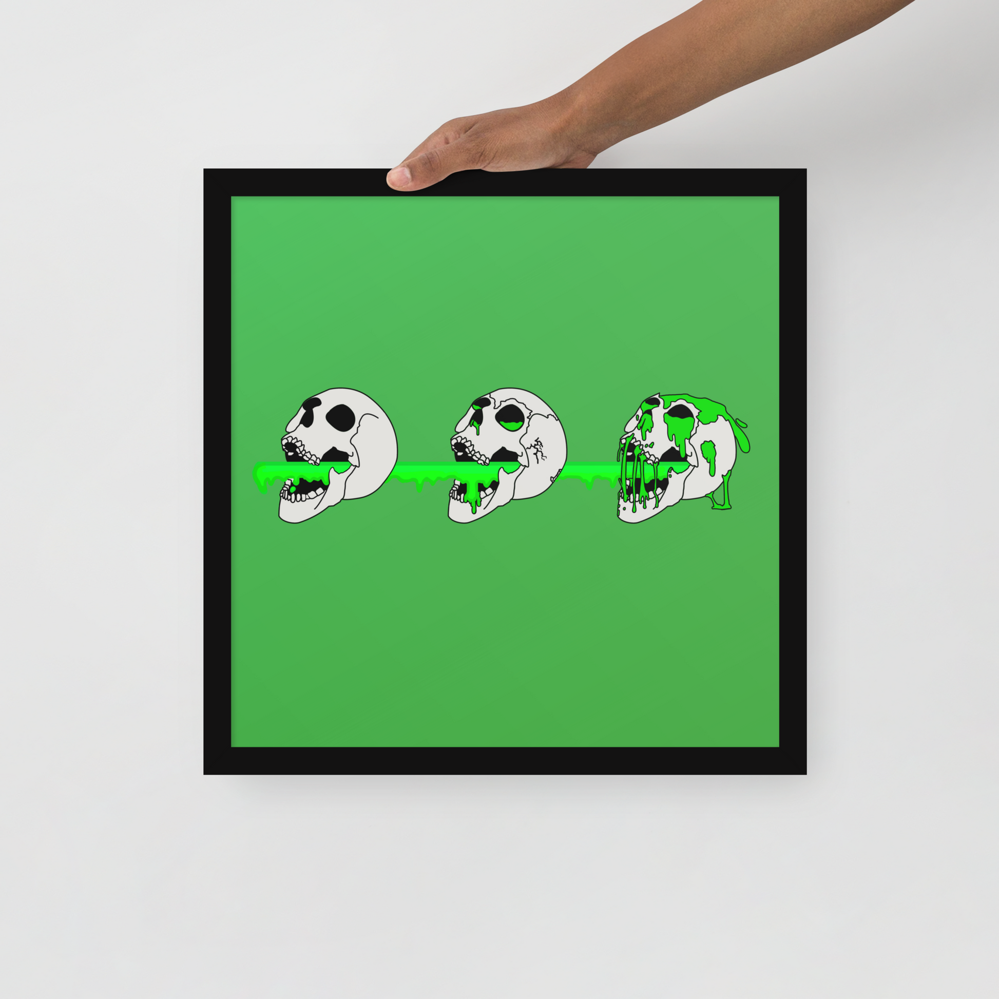Framed artwork with a green background and black frame, featuring three cartoonish skull designs.