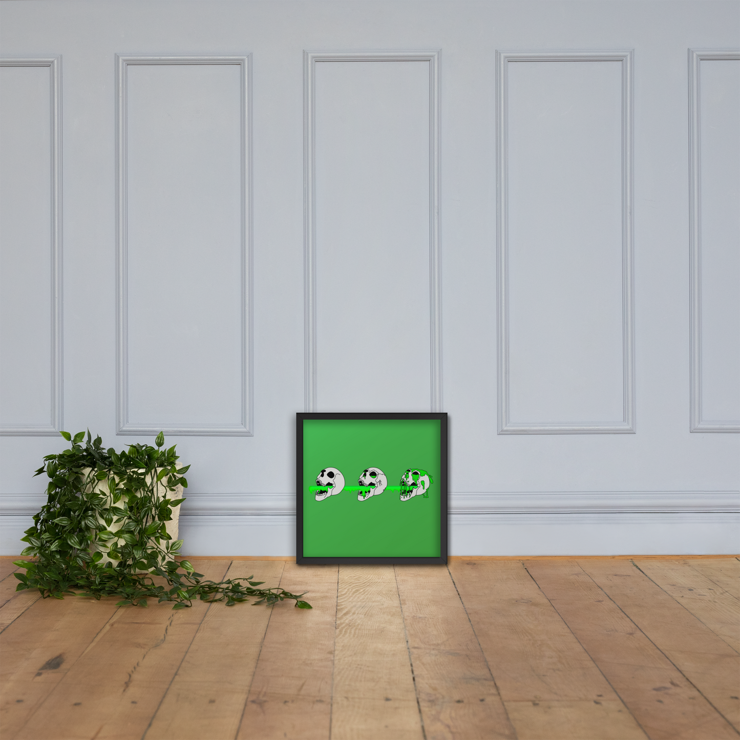 Framed artwork with a green background and 3 skulls shooting green slime from their mouths with a gradient background in a horror style design on a wooden floor against a white paneled wall by a plant.