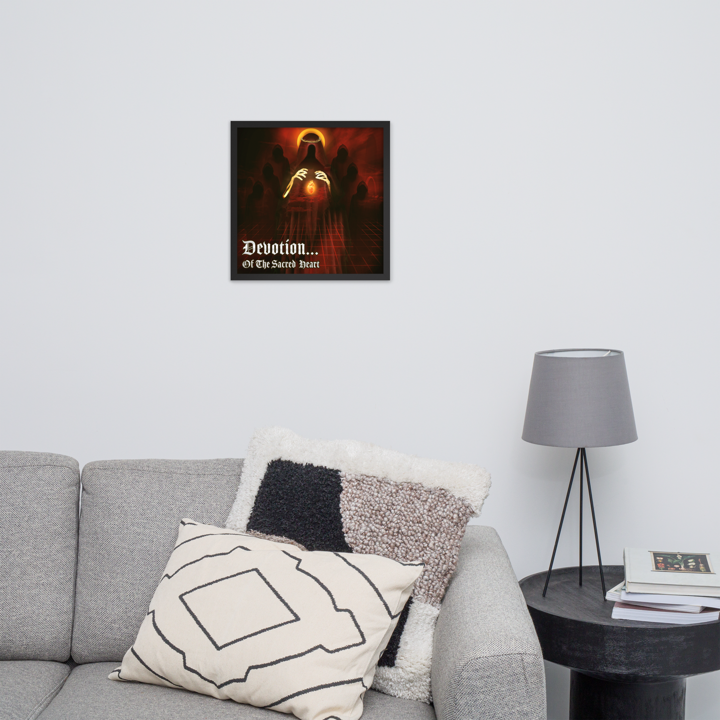 Framed artwork titled 'Devotion... Of The Sacred Heart' above a couch in a living room area with a lamp and books