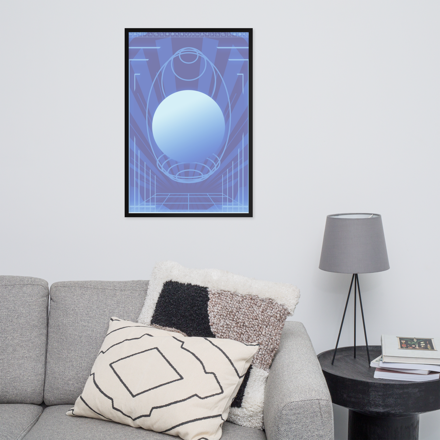 A framed abstract art print featuring a blue and light blue geometric design with a central oval, displayed on a white wall above a gray sofa with decorative pillows and next to a black side table with a lamp.