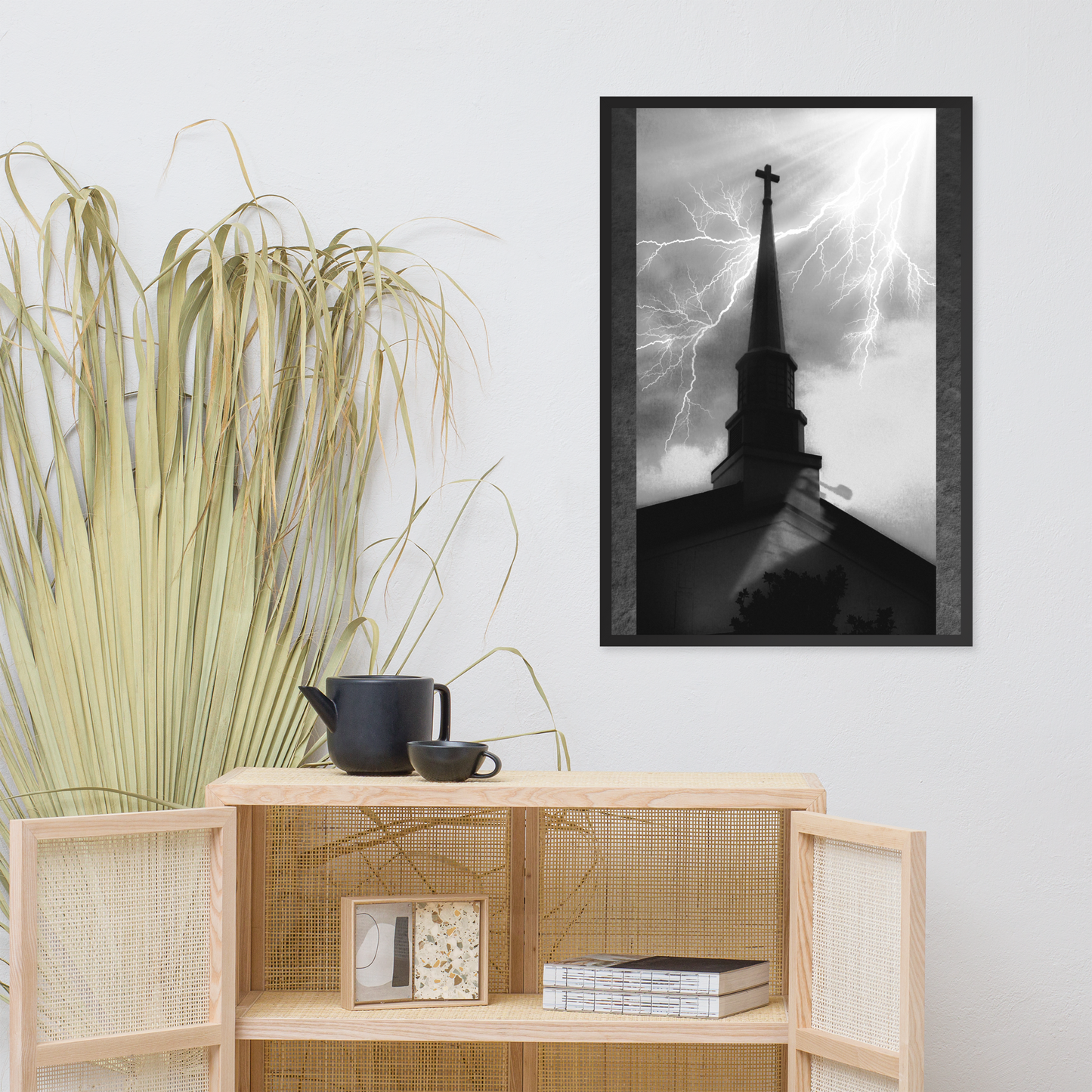 A framed black and white art print of a church steeple with lightning, displayed on a light gray wall above a wooden shelf with decorative items.