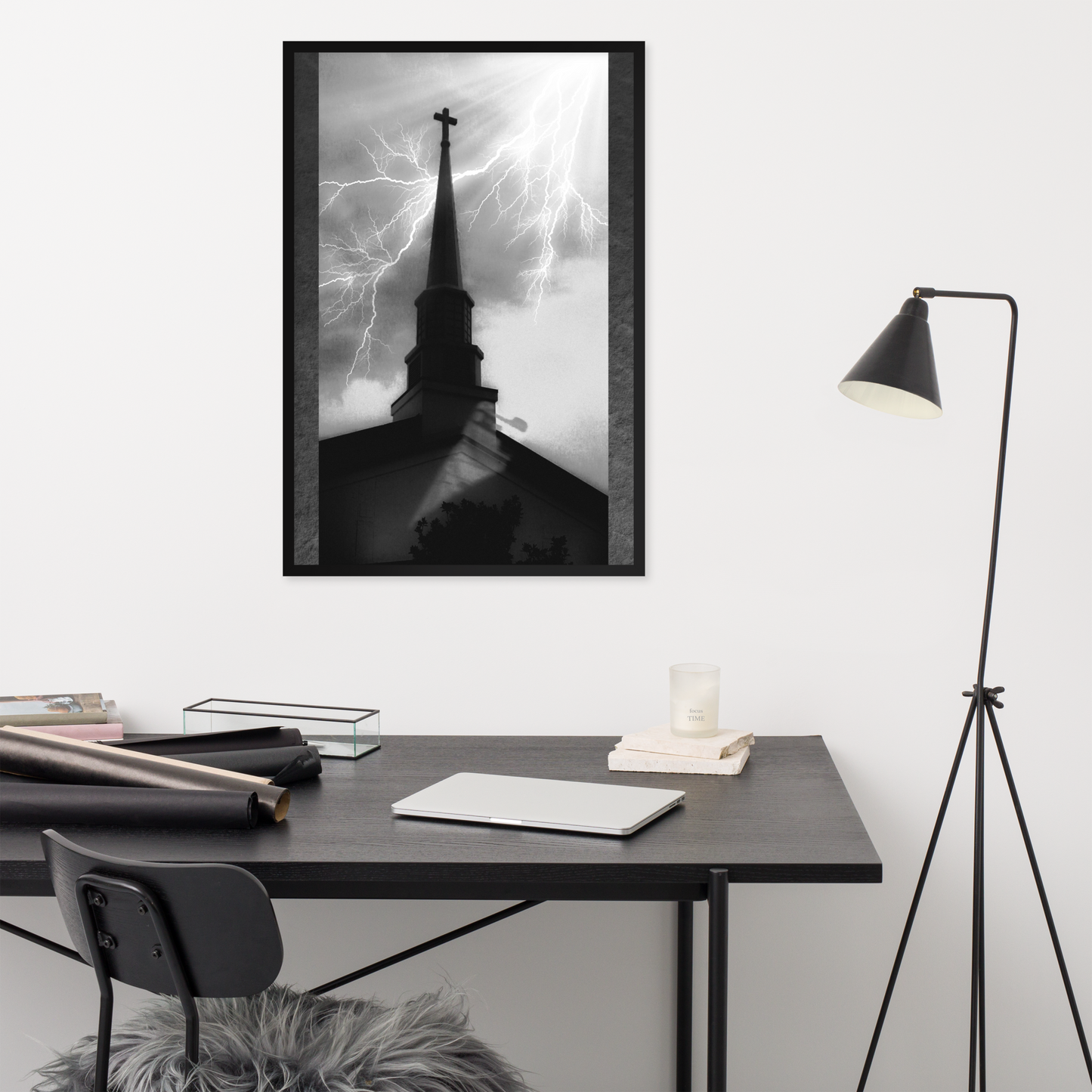 A black-framed black and white art print of a church steeple under lightning storm clouds, displayed on a white wall above a modern desk setup with a chair and floor lamp.