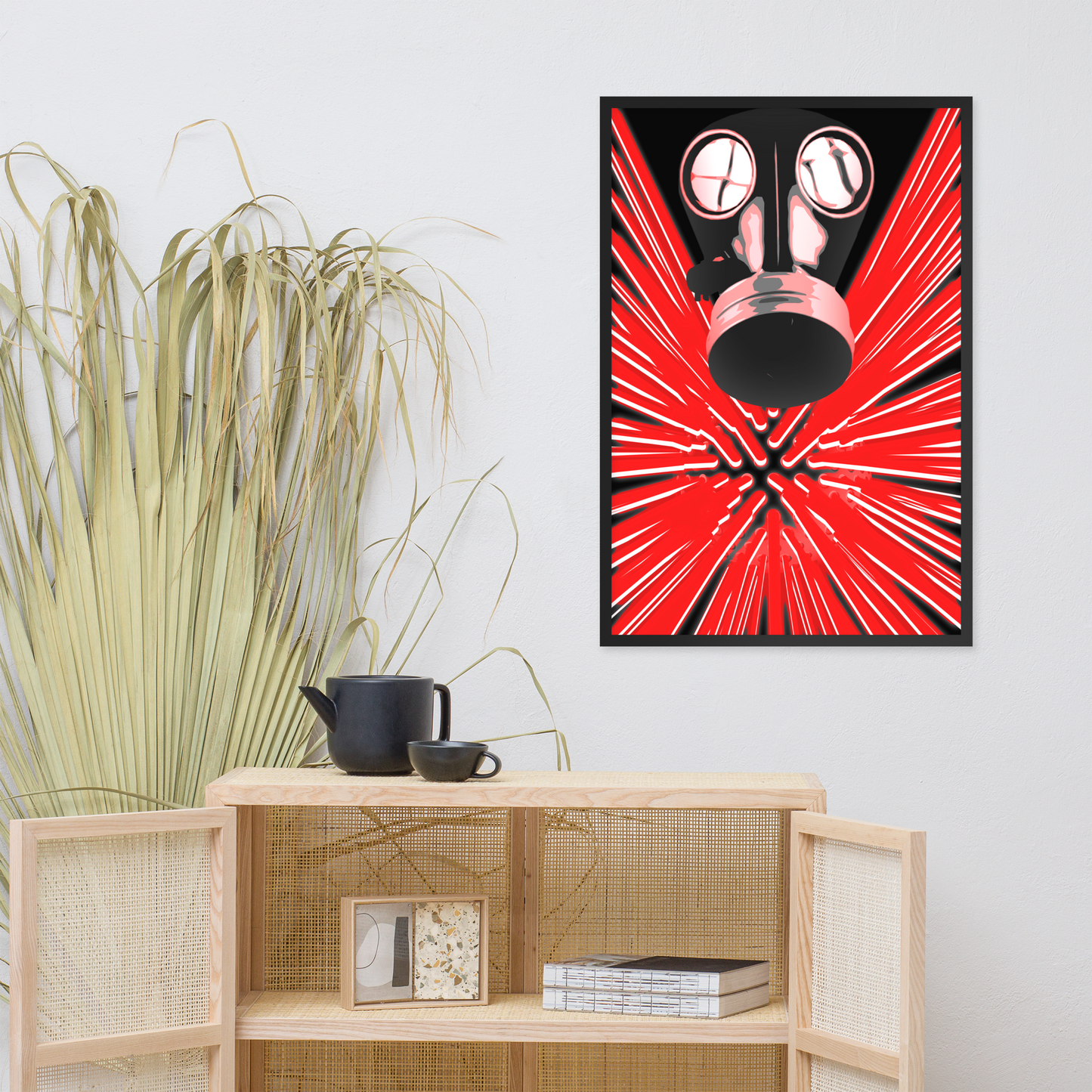 A framed wall art print featuring a black gas mask over a bright red burst pattern, displayed above a light wood cabinet in a styled interior with a potted plant and small decor items.