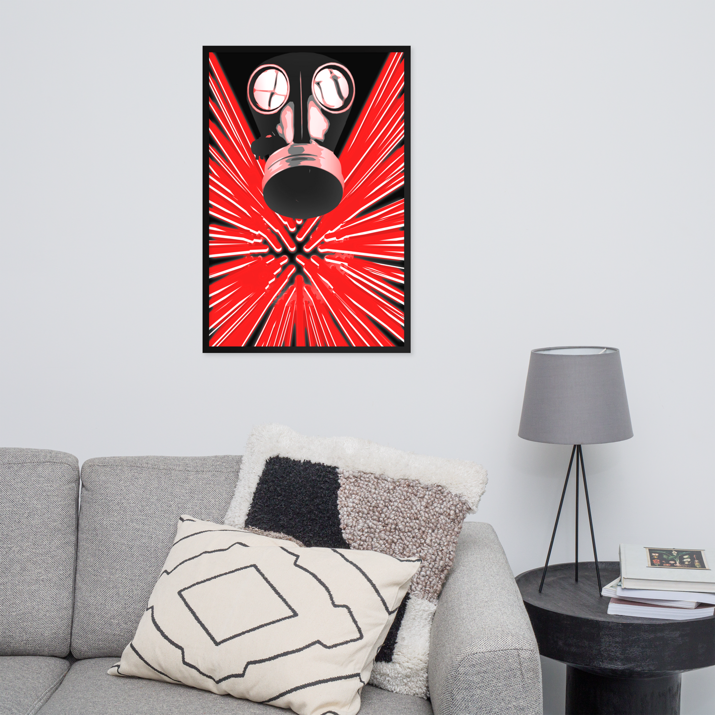 A framed wall art print featuring a stylized black gas mask over a bright red burst pattern, displayed above a gray sofa in a modern living room setting with pillows, a side table, and a lamp.